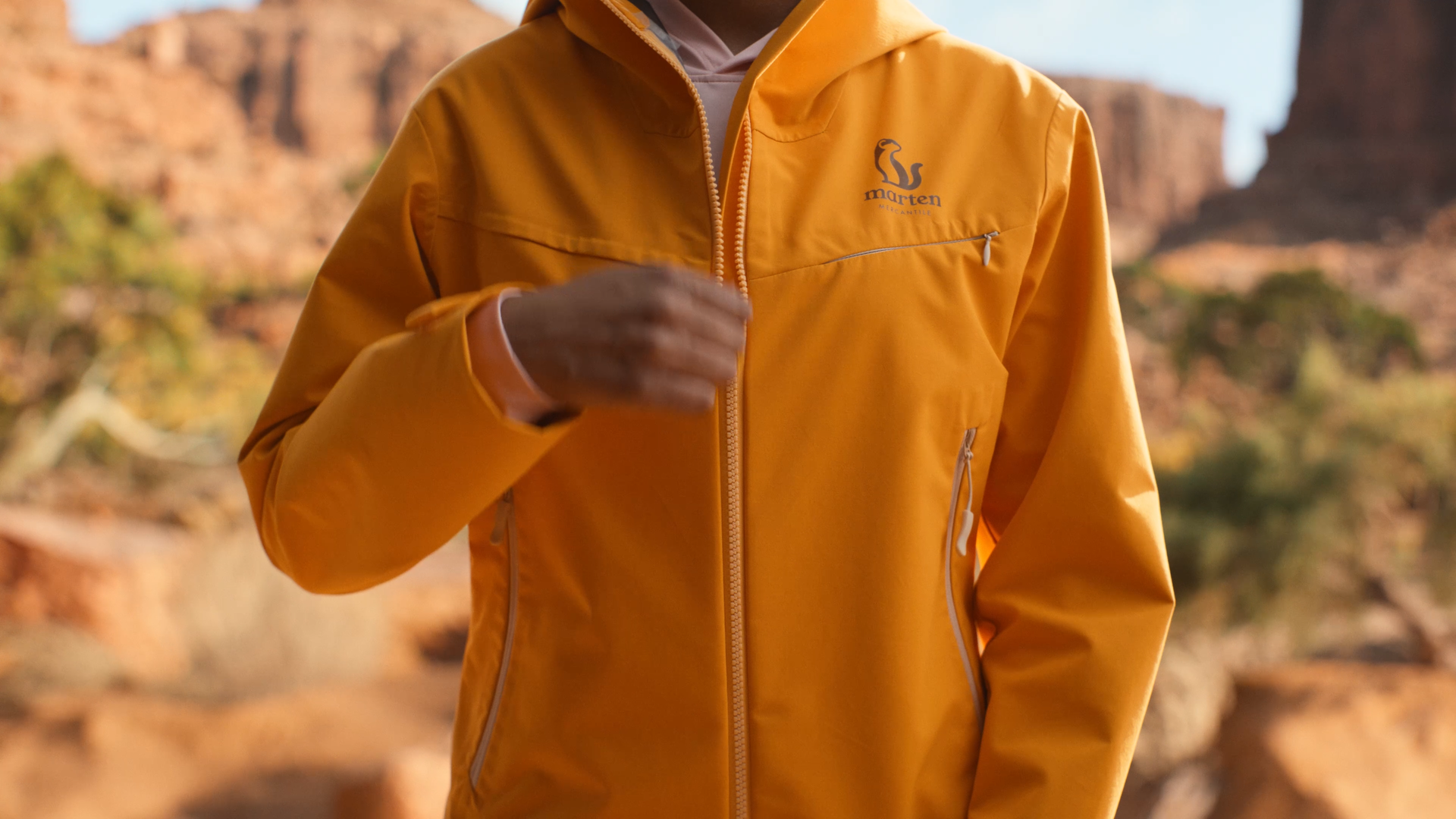 Person wearing a yellow Marten outdoor jacket with front zipper, standing outdoors with desert landscape and rock formations in the background.
