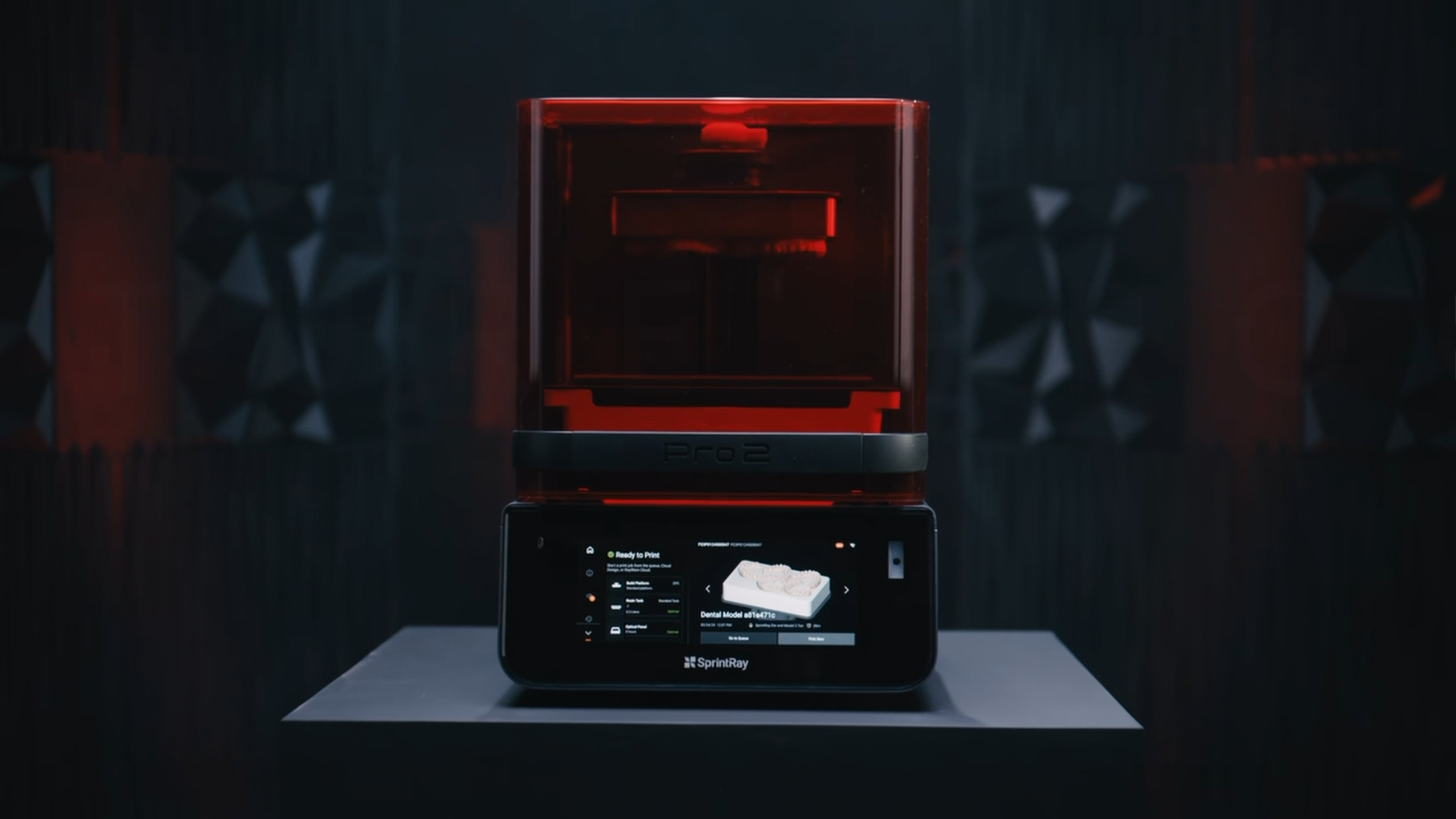 A 3D printer with a red casing is printing a dental model. The printer is labeled 'Pro2' and is placed on a table. In front of the printer, there is a digital screen displaying print status information.