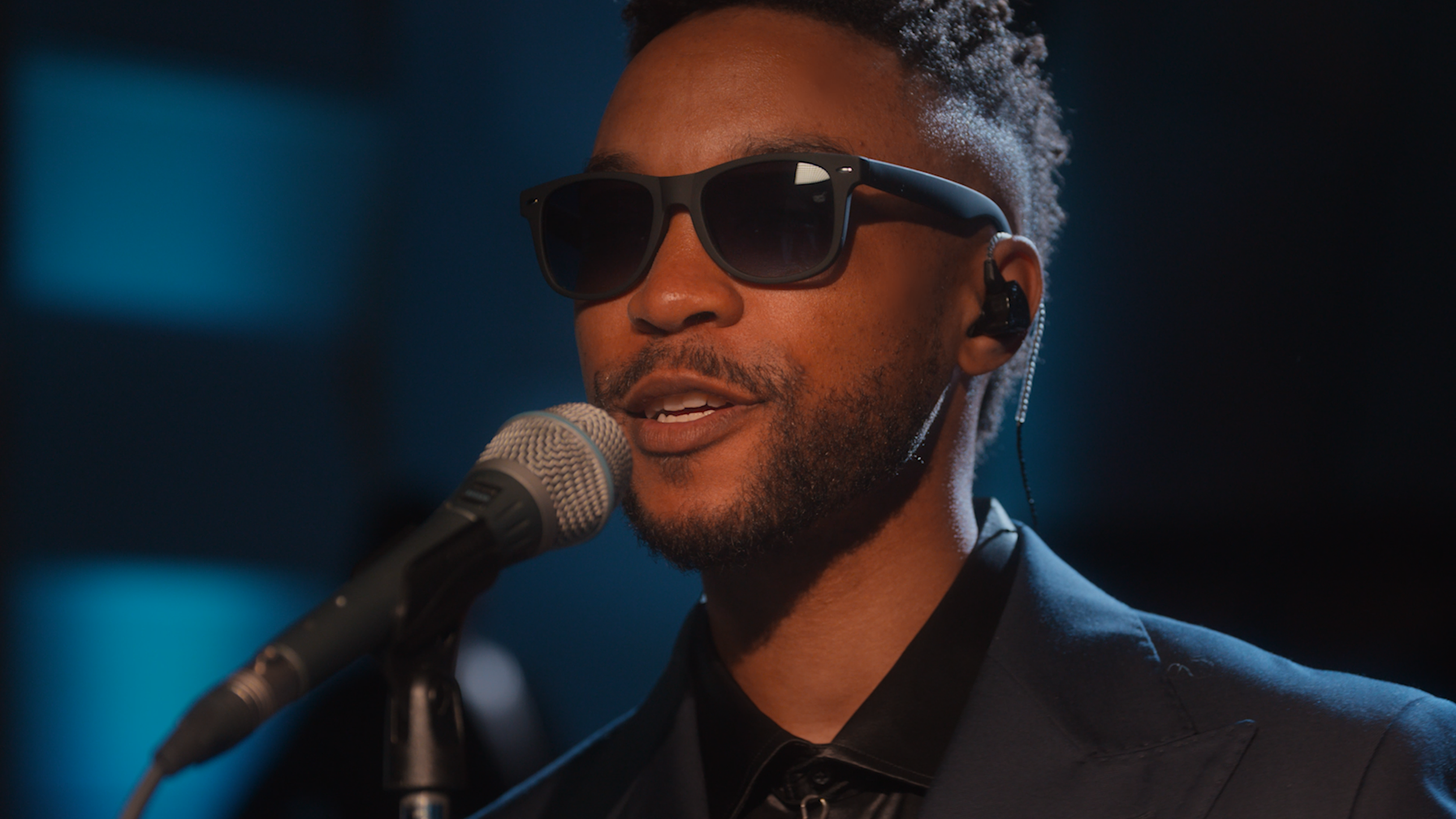 A man wearing dark sunglasses and an in-ear monitor is speaking into a microphone, with a dark blurred background.