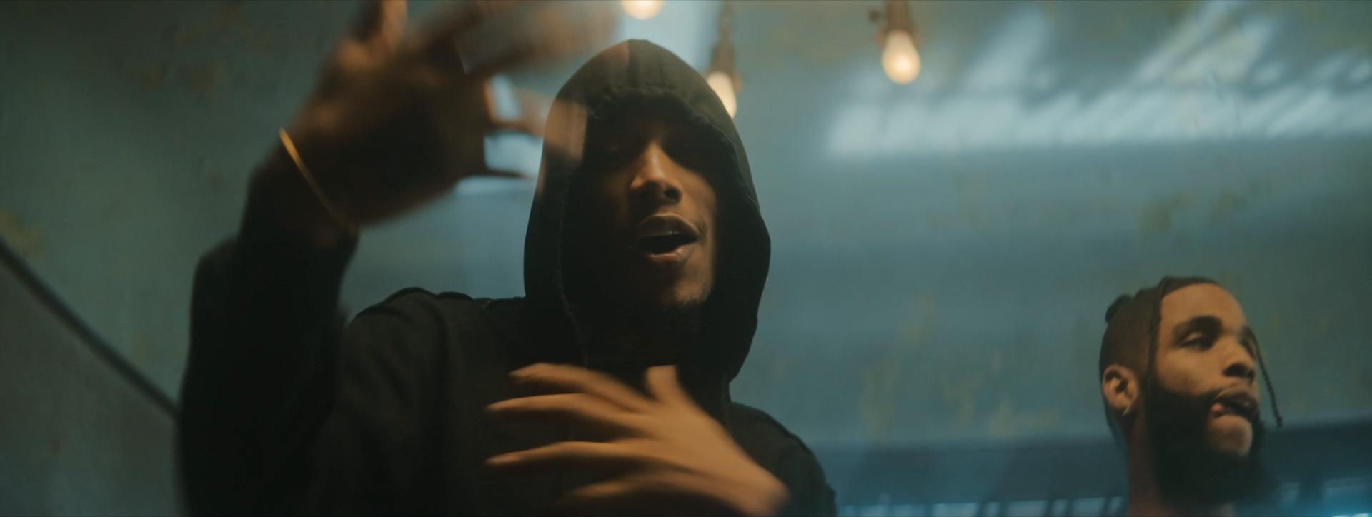 Two men in hooded sweatshirts in a dimly lit room with hanging lights, one of them gesturing with his hand.