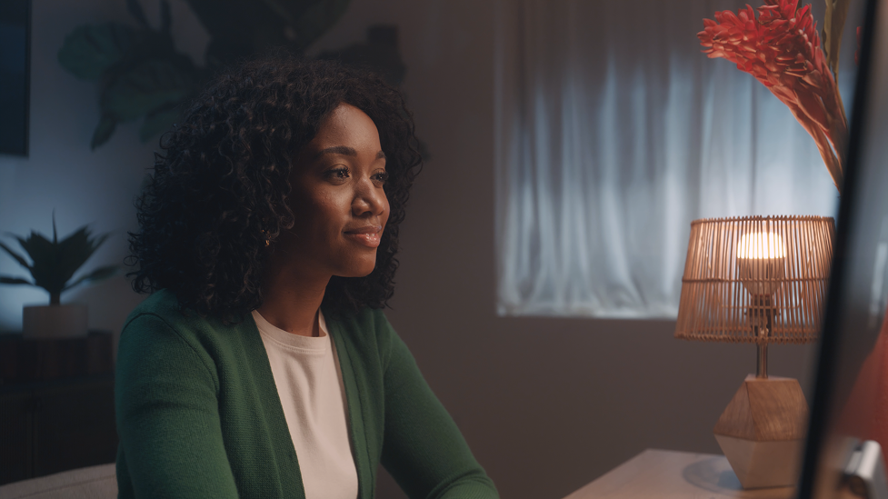 A woman with curly hair sitting at a desk, looking at a computer screen, in a softly lit room with a table lamp and plants.