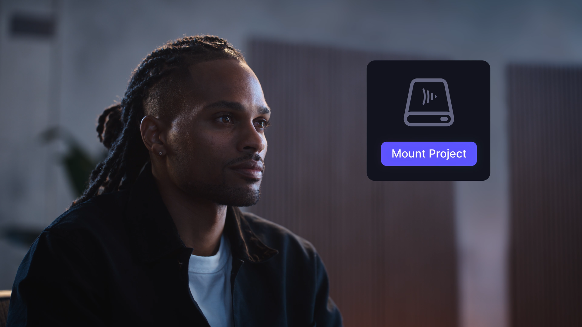 A man with dreadlocks and a dark jacket looking attentively at a holographic computer interface that displays a storage drive icon and a 'Mount Project' button.