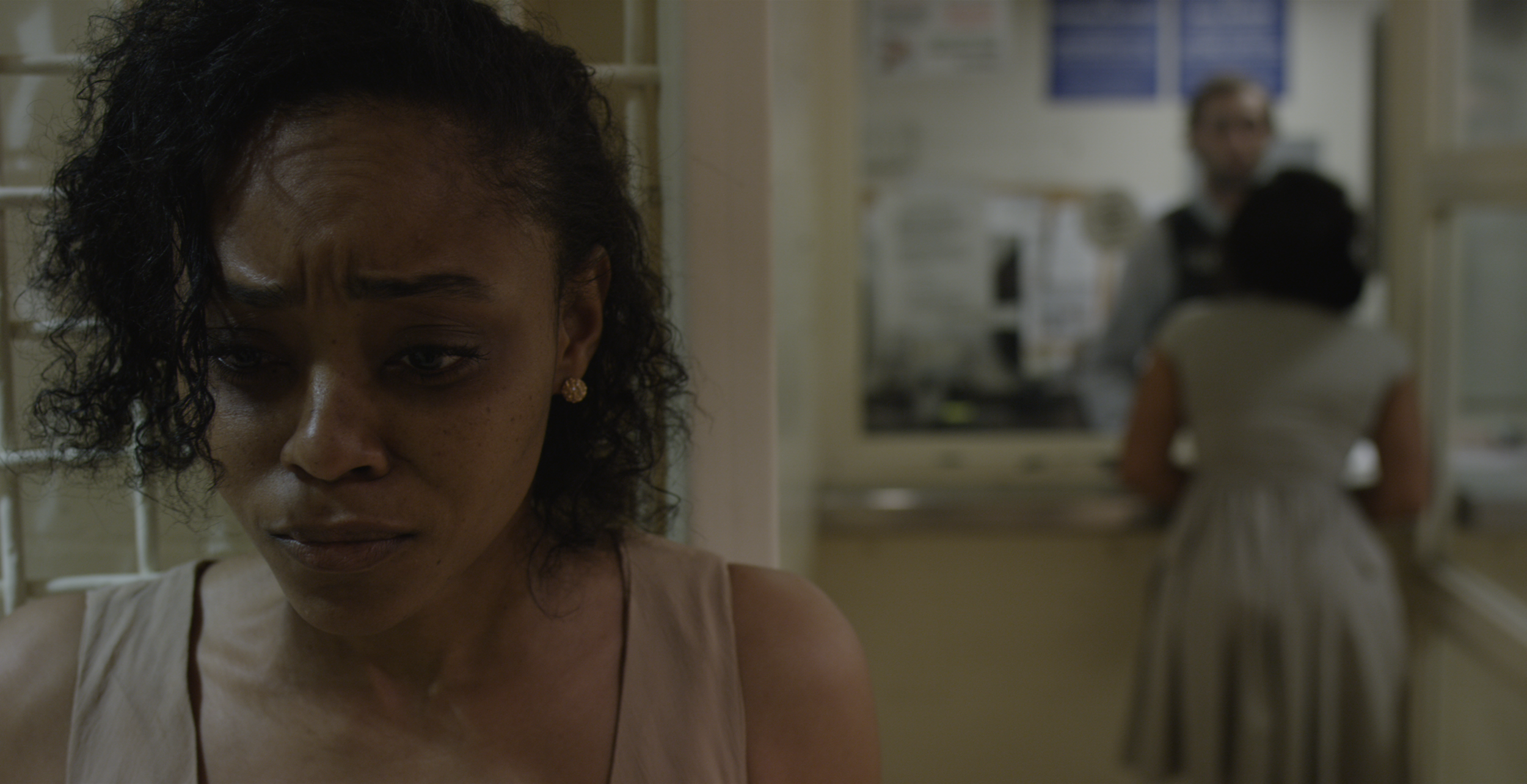 A young woman with curly hair and earrings looks down, visible from the shoulders up, with a blurred background of a room with a person standing and examining something.