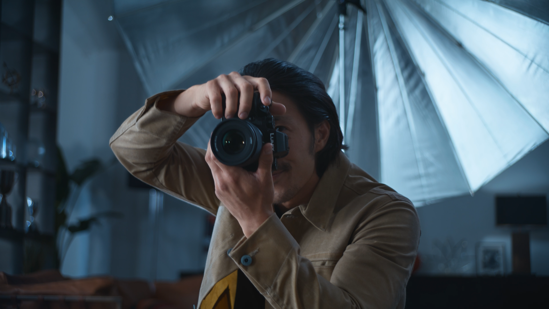 Person with dark hair taking a photograph with a camera indoors, with a large reflective umbrella overhead and a blurred background.