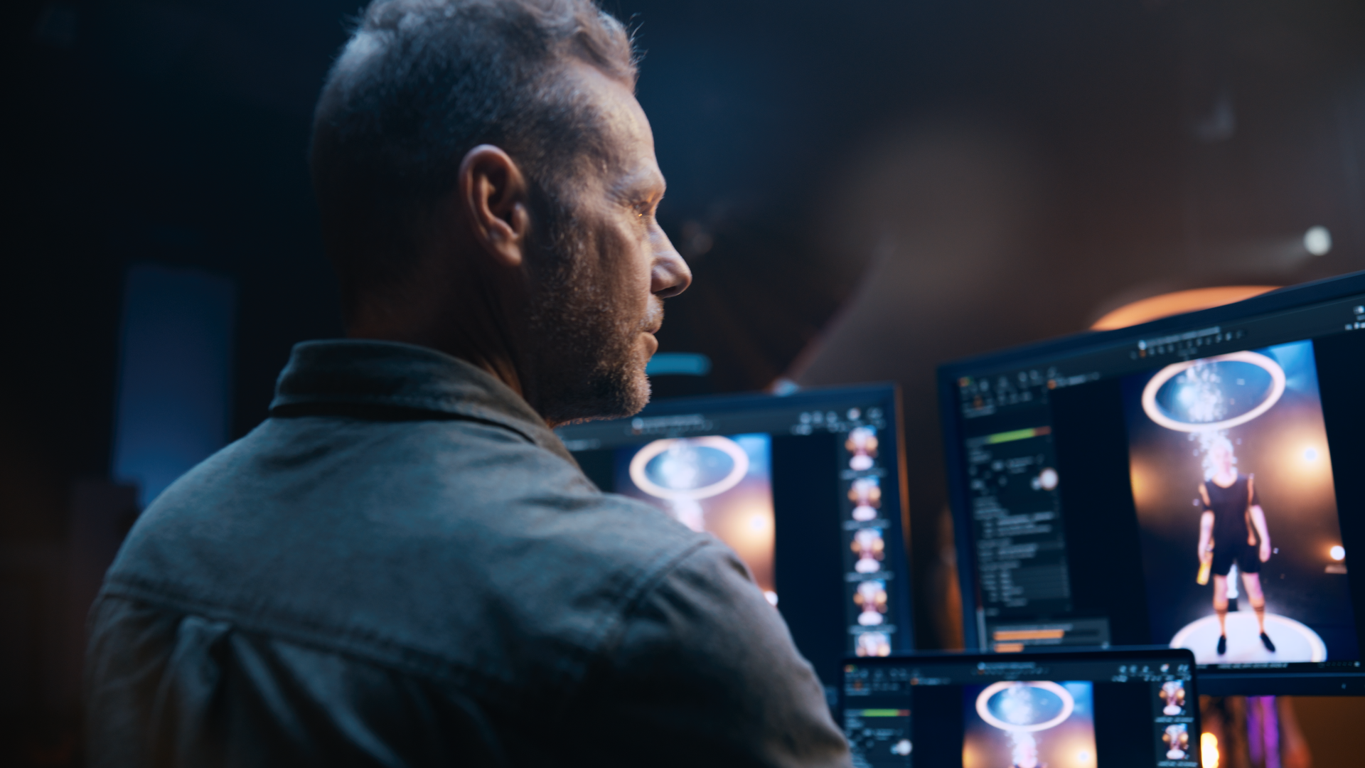 A man with short, styled hair and a beard working on a computer in a dark room. The screens display an animated character and digital editing software.