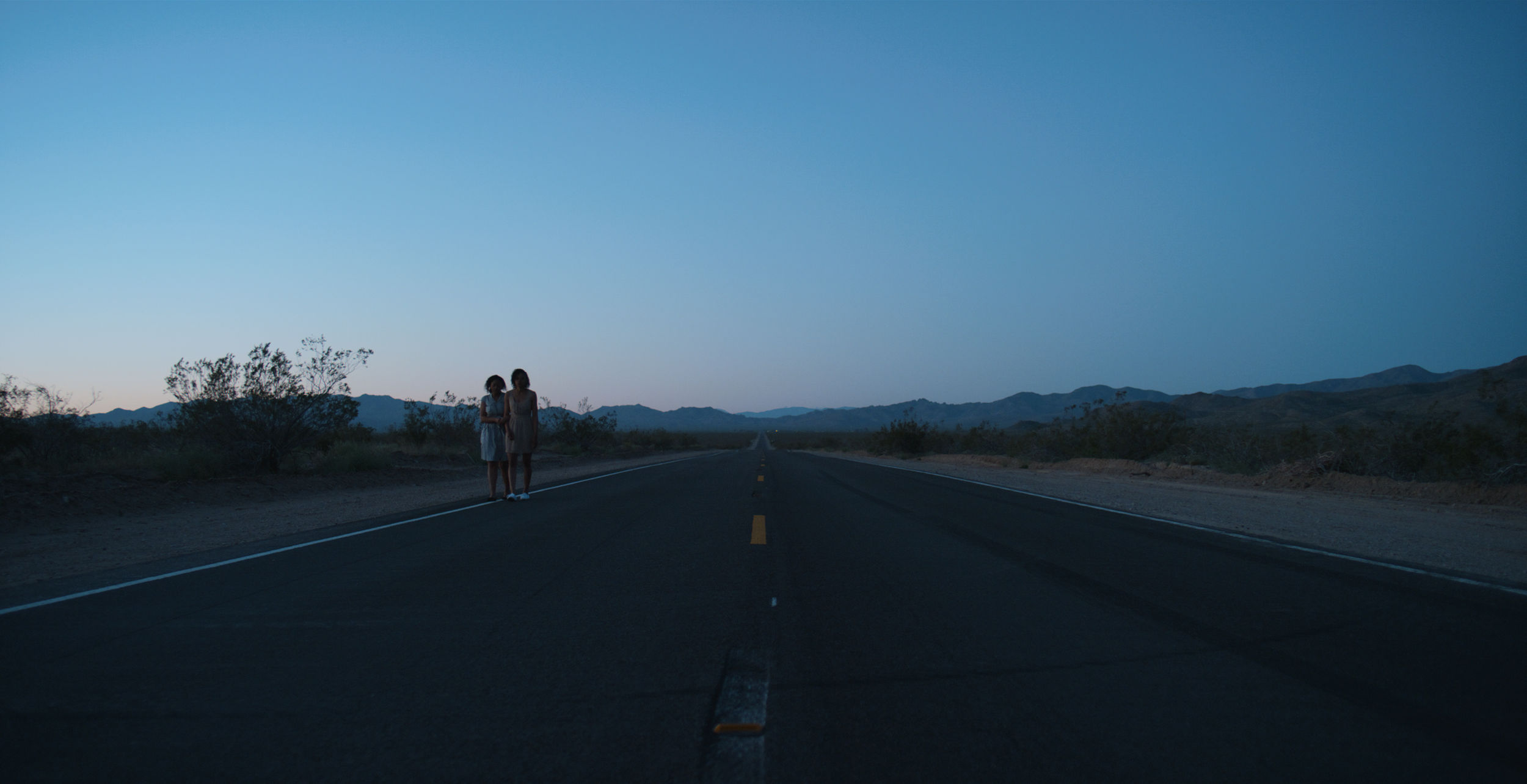 Two women stand on a deserted road in a desert landscape during dusk, with mountains in the background.