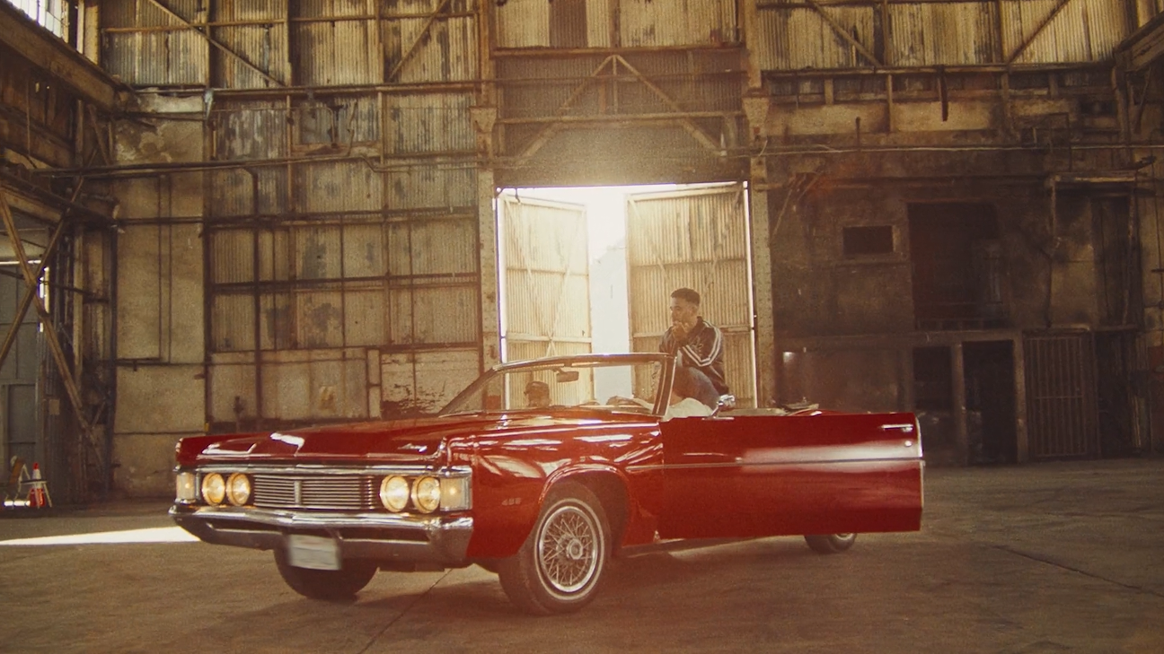 A young man in casual clothing sitting on the edge of a shiny red convertible car inside a large, rustic warehouse with wide-open doors backlit by bright sunlight.