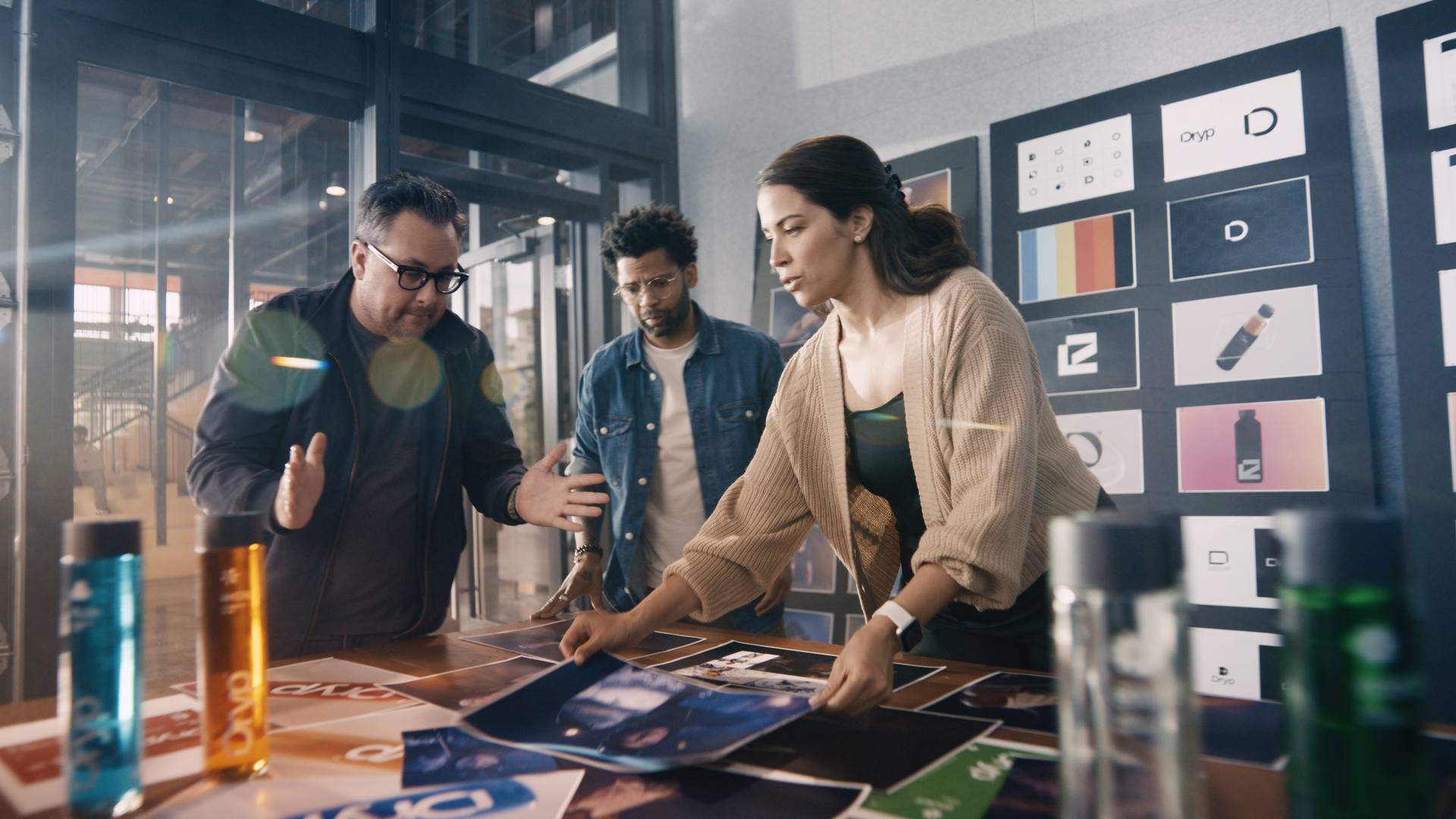 Three people standing around a table, discussing printed materials in a modern office setting, with design sketches on the wall and bottles of paint or ink in the foreground.