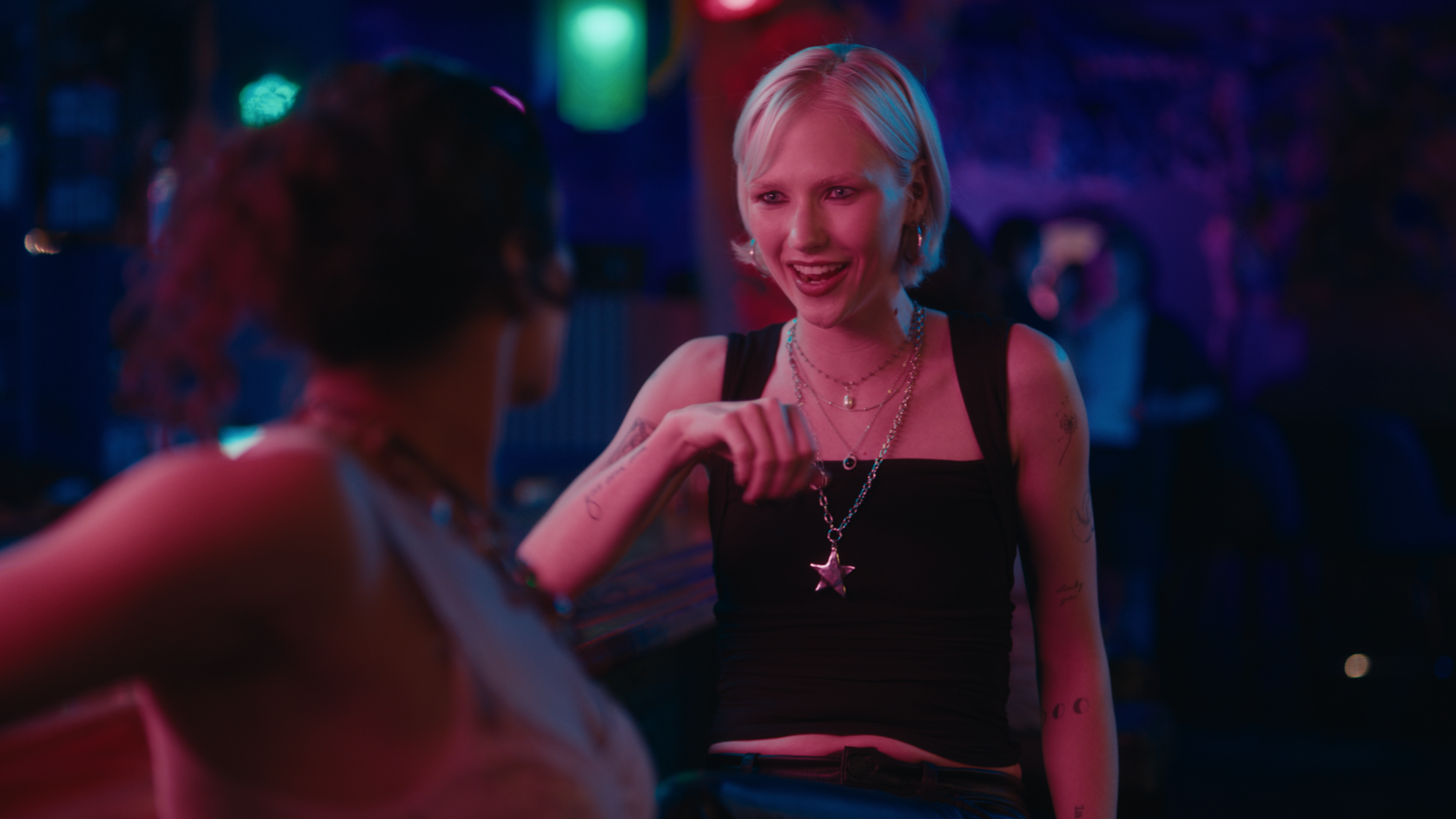 A woman with short platinum blonde hair, wearing a black tank top, layered necklaces, and star-shaped pendant, smiling and talking with another woman in a dimly lit, colorful nightclub or party setting.