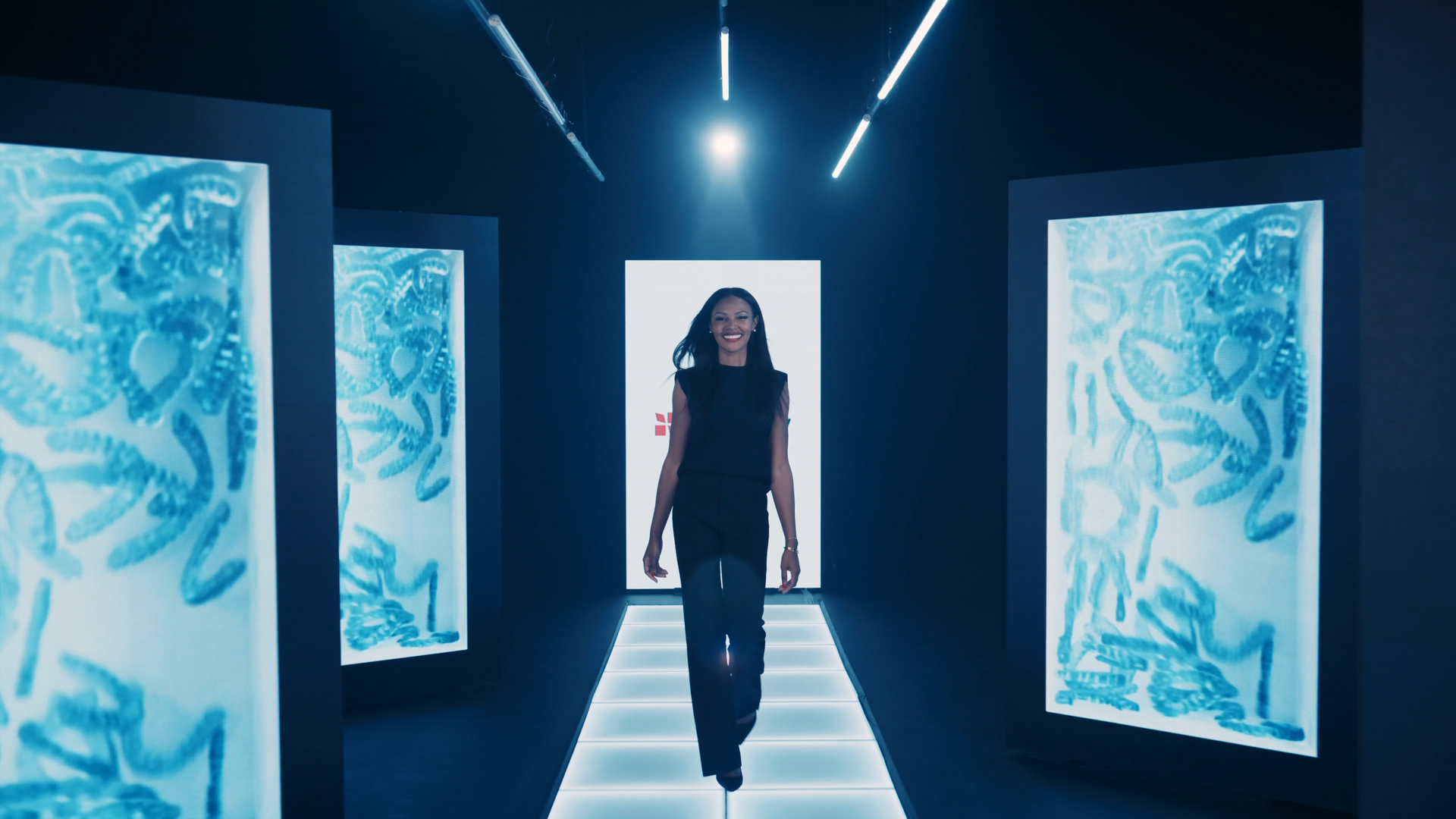 A woman in black pants and sleeveless black top walking in a futuristic, dark corridor surrounded by illuminated digital screens featuring abstract blue designs.