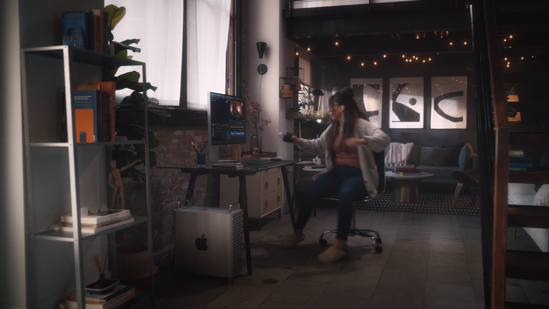 A woman in a cozy, modern living space, sitting on a rolling office chair, holding a cup and dancing with a lively expression. The room features a brick accent wall, a large window with sheer curtains, a desk with a computer, and artwork on the wall.
