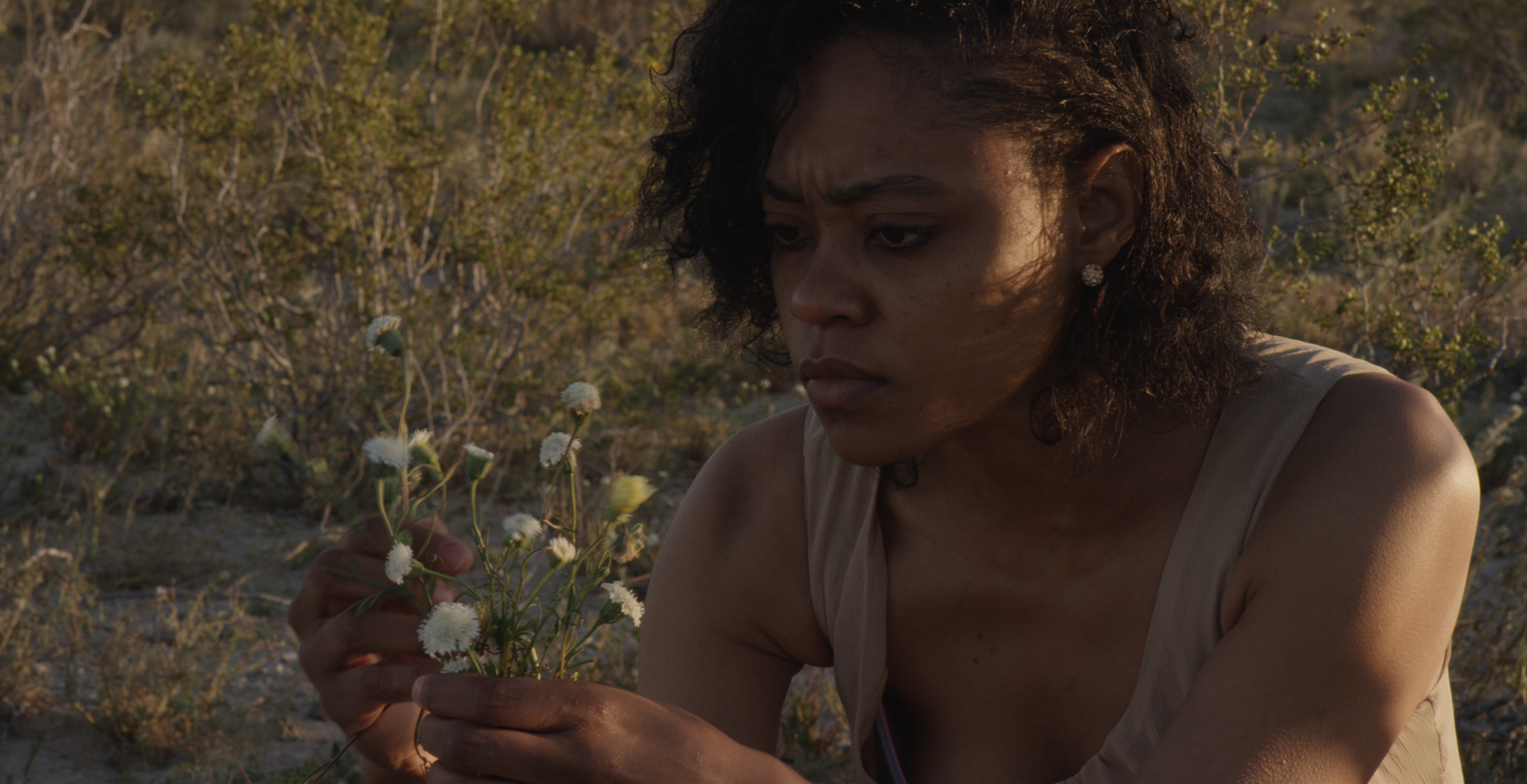 A woman with curly hair and earrings is sitting outside in a desert-like area with shrubs, holding a small bouquet of white flowers and examining them closely.