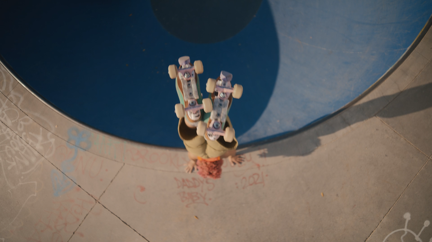 A person with curly pink hair standing on a skateboard at a skate park, viewed from above, with graffiti on the concrete.