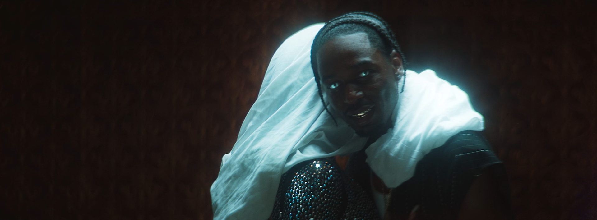 A man with braided hair wearing a black shirt with sequins and a white garment draped over his shoulder, looking into the camera with a dark background.