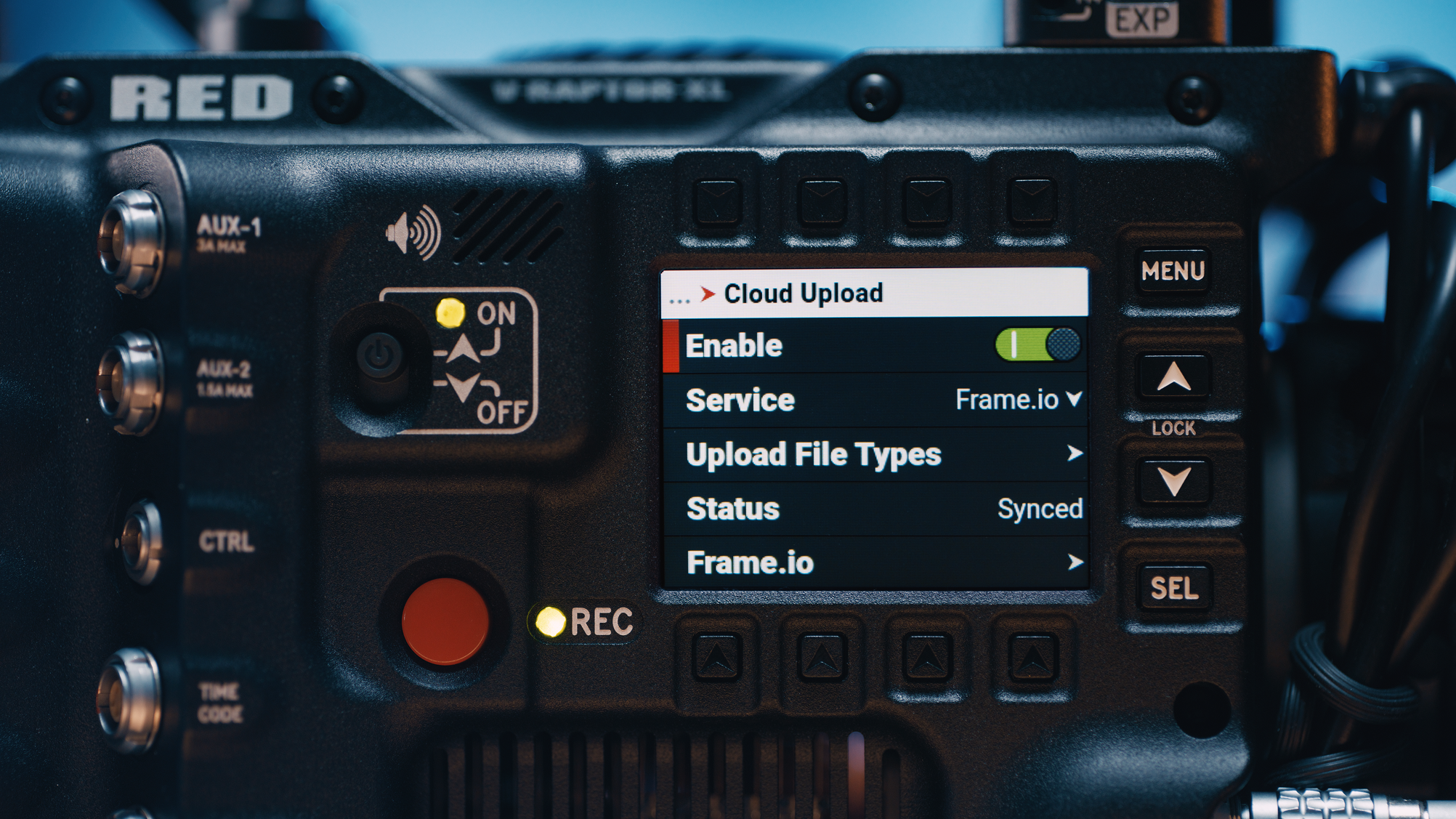 Close-up of a camera's screen displaying menu options for cloud upload, enabling, service, upload file types, status, and frame.io. The camera has various buttons and dials, including a red record button and an AUX input.