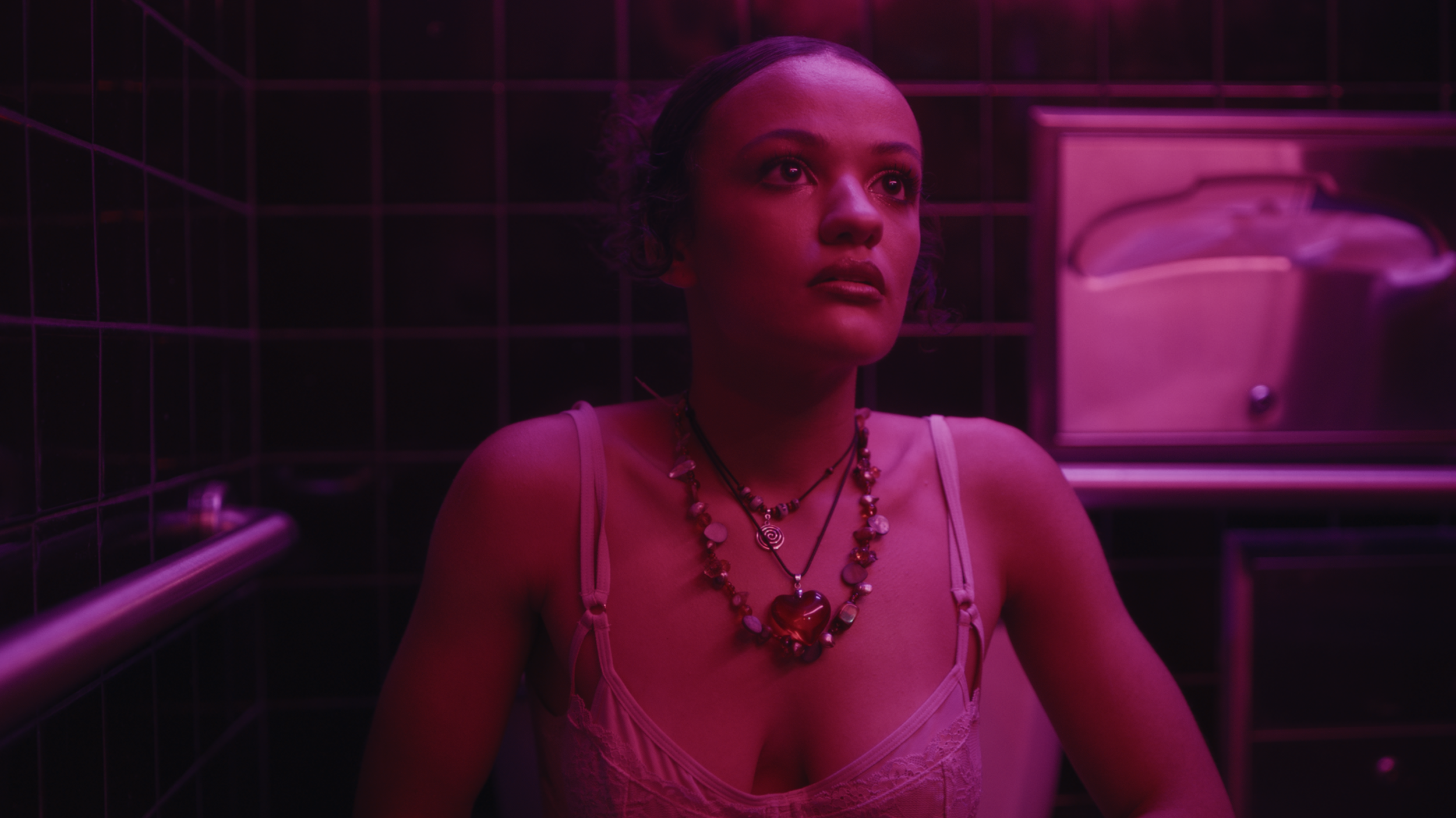Woman with multiple necklaces, wearing a pink top, in a dimly lit room with black tiled walls and purple lighting.