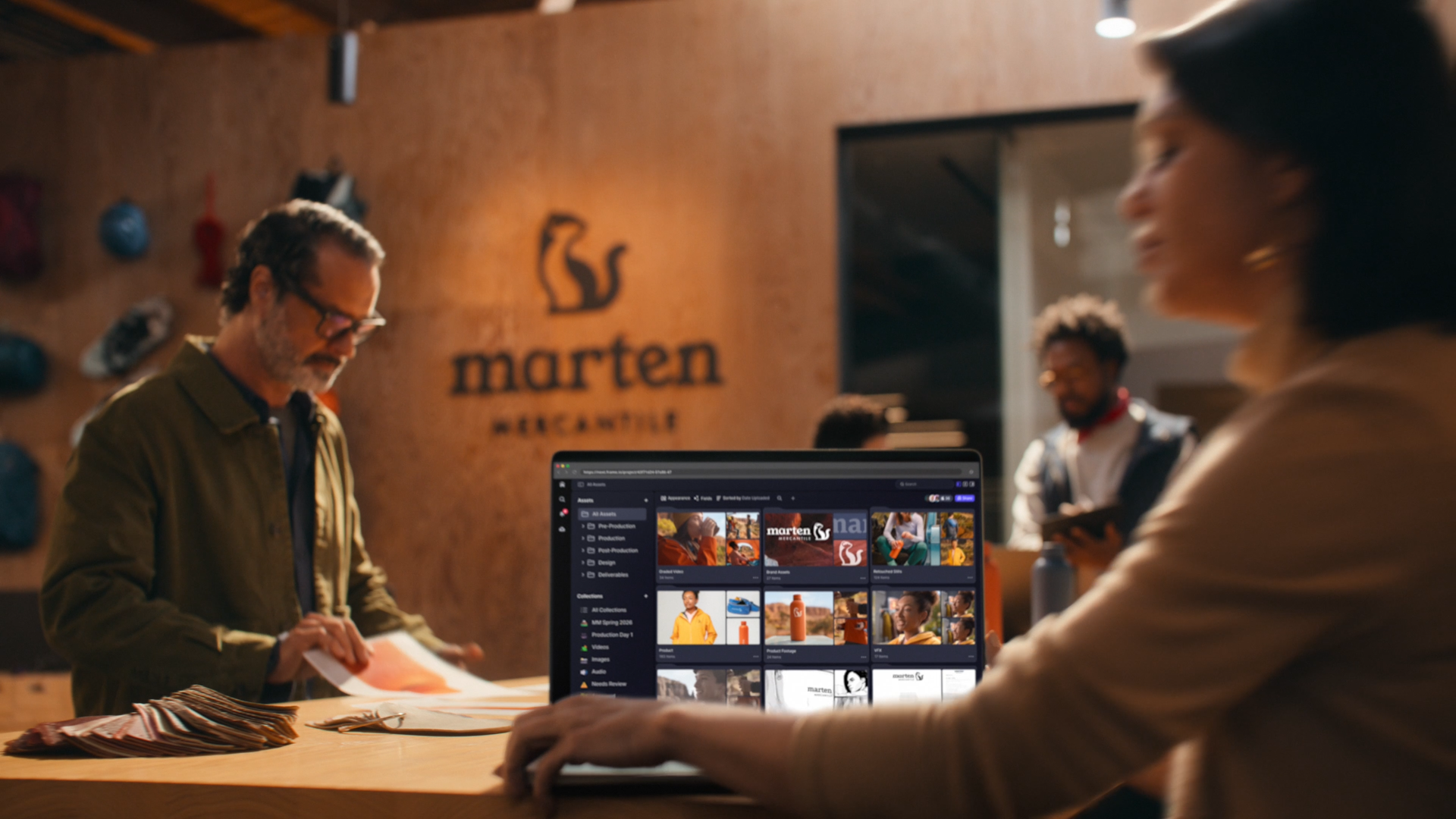 People working in a creative studio space with a sign that says 'marten MERCHANTILE'. A laptop with a photo editing or asset management software open shows images of people, products, and branding elements.