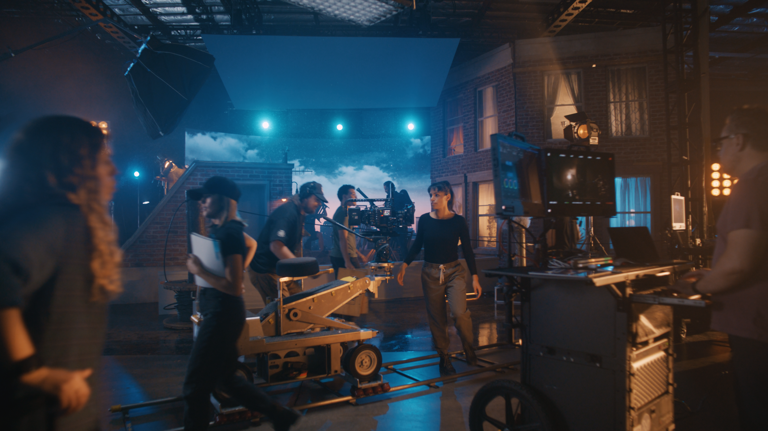 Film crew working on set with camera and lighting equipment on a rooftop at night, with a cloudy sky backdrop, bricks, and windows in the background.