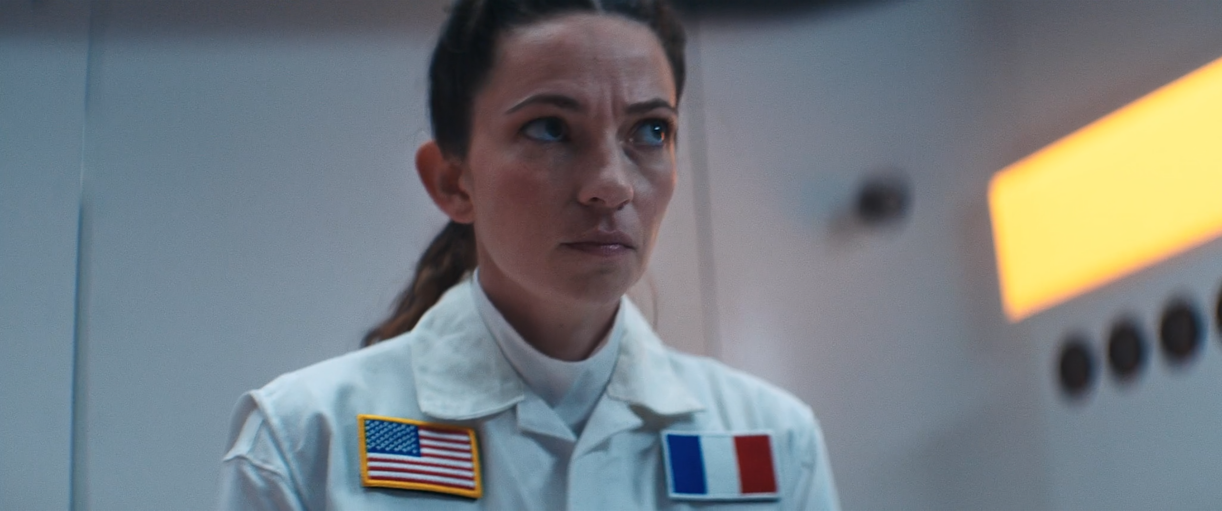 A woman dressed as an astronaut or space crew member, wearing a white uniform with American and French flags on her chest, looking intently at a screen or control panel in a plain, light-colored room.