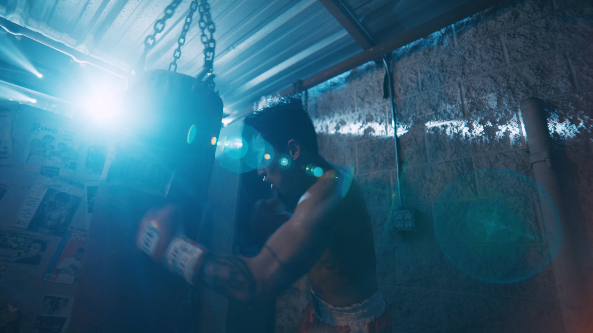 A young man is playing with a punching bag in a garage or basement with concrete walls. Bright sunlight is shining in from outside, creating lens flare effects in the image.