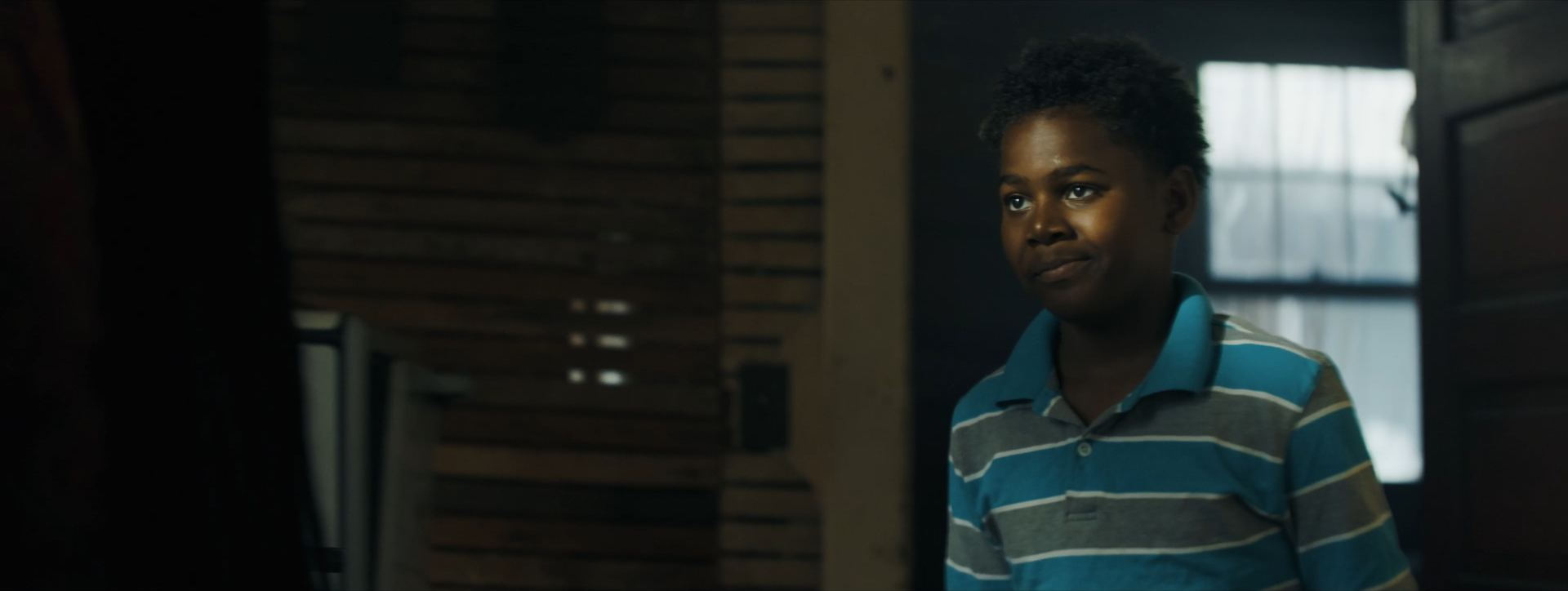 A young boy with dark curly hair wearing a blue, gray, and white striped polo shirt standing indoors near a wooden wall, with a window in the background.