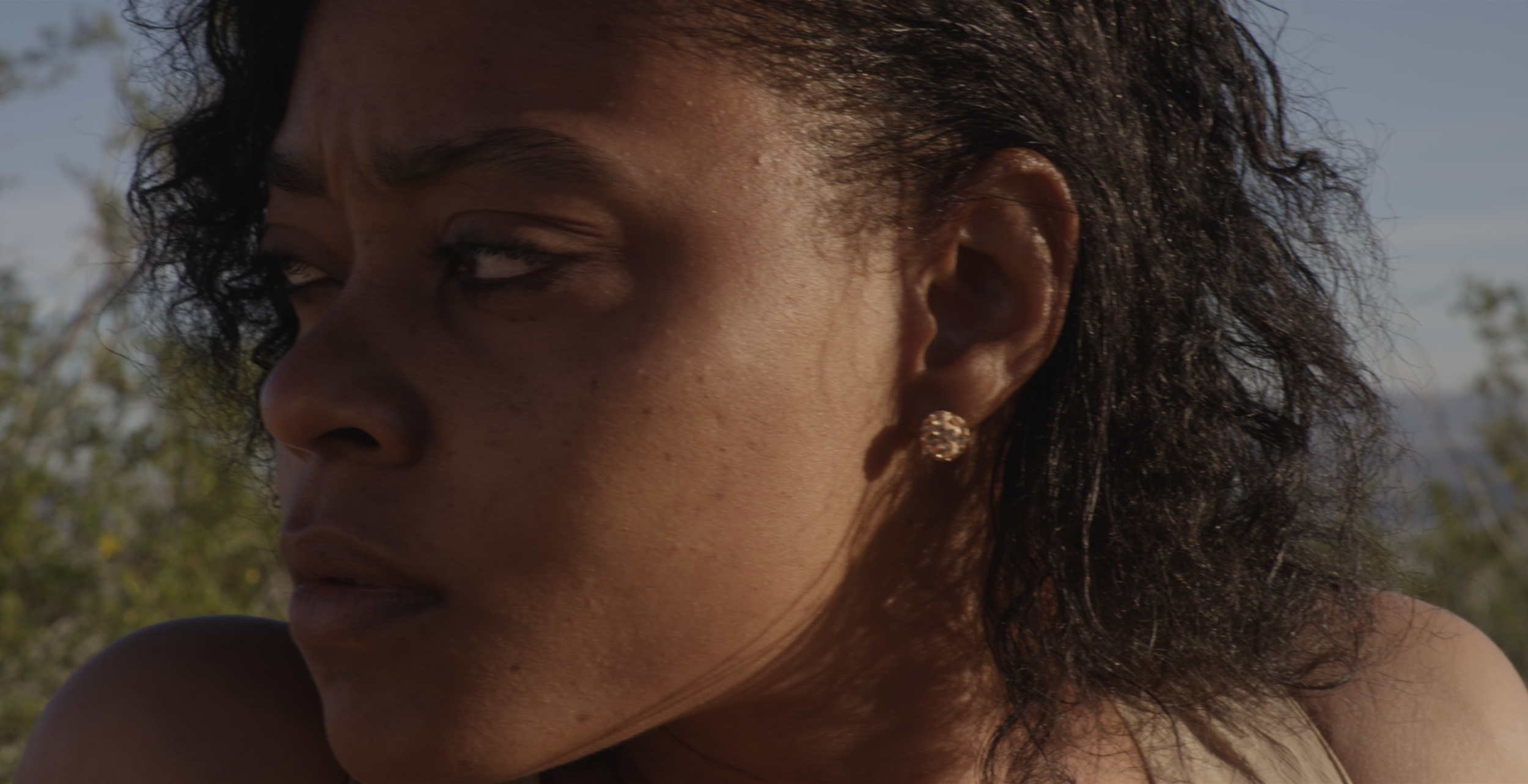 Close-up of a woman's face, showing her eye, cheek, and ear, with short, curly dark hair and a sparkling earring, outdoors with trees in the background.