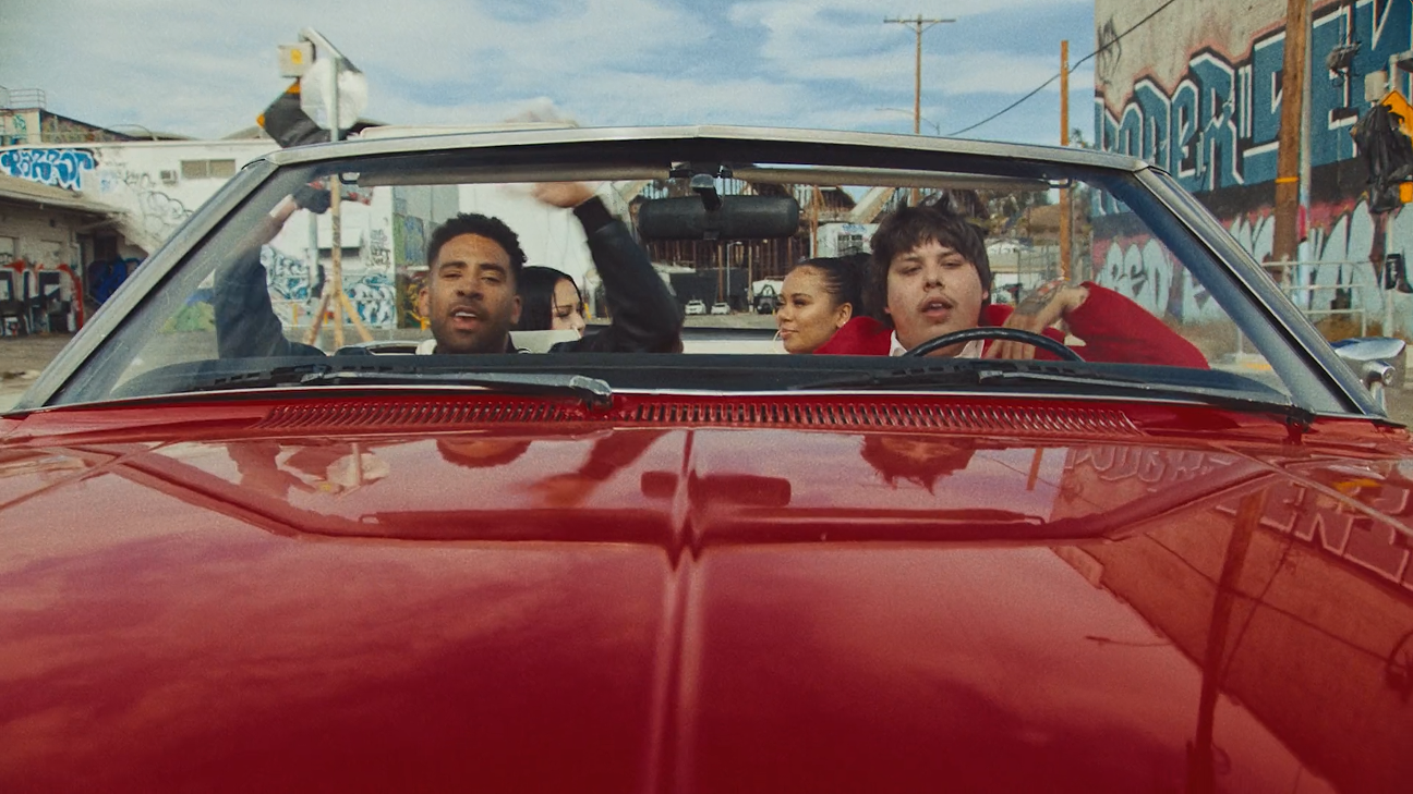 A red convertible car with four young people inside, driving through an urban area with graffiti-covered walls and utility poles.