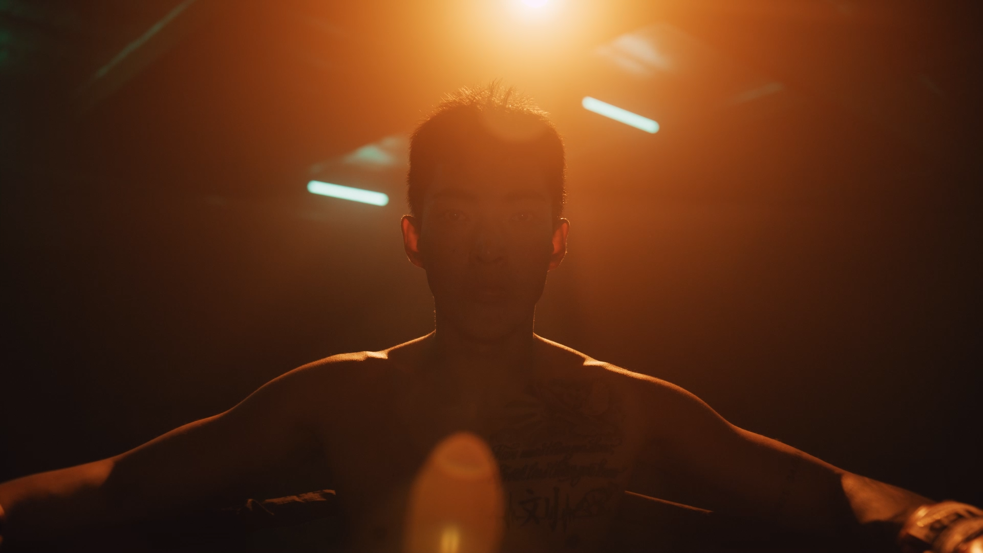 A shirtless man with tattoos on his chest and arm, standing in a dimly lit environment with warm orange light overhead, looking directly at the camera.