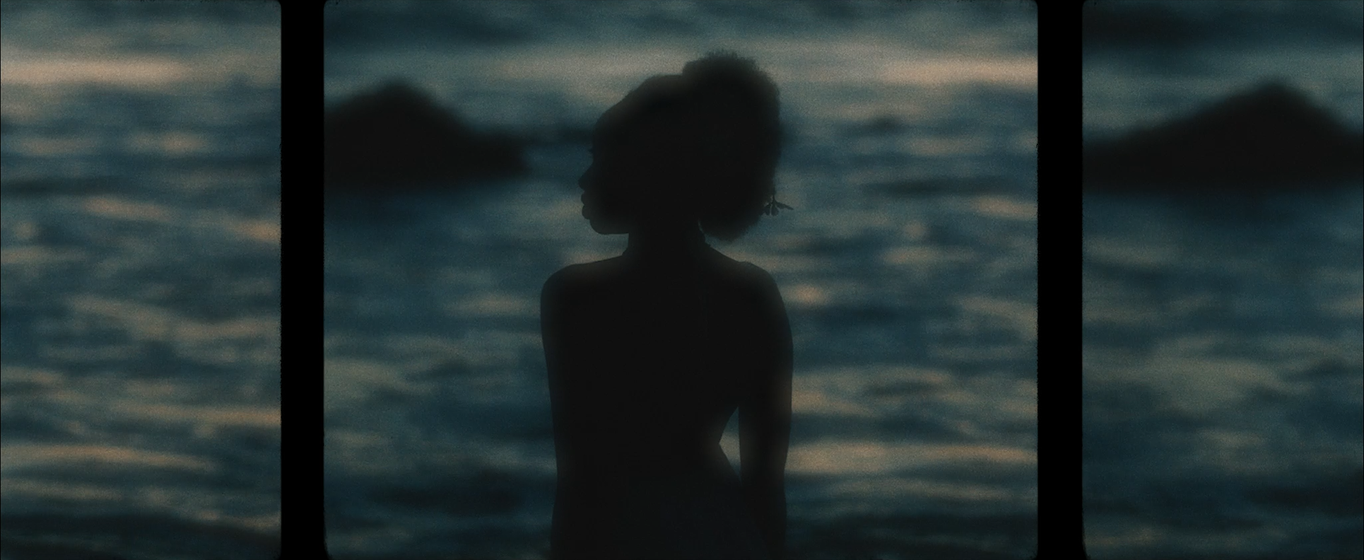 Silhouette of a woman with natural curly hair standing in front of a body of water during sunset or dusk, with reflections on the water.