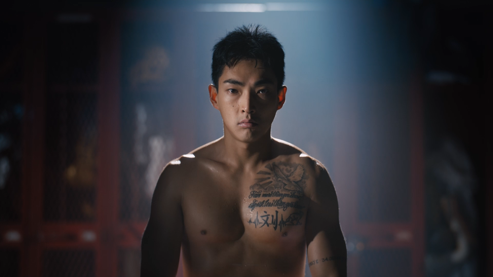 A shirtless young man with tattoos on his chest and shoulder looks directly at the camera, standing in a dimly lit room with a dramatic spotlight focused on him.