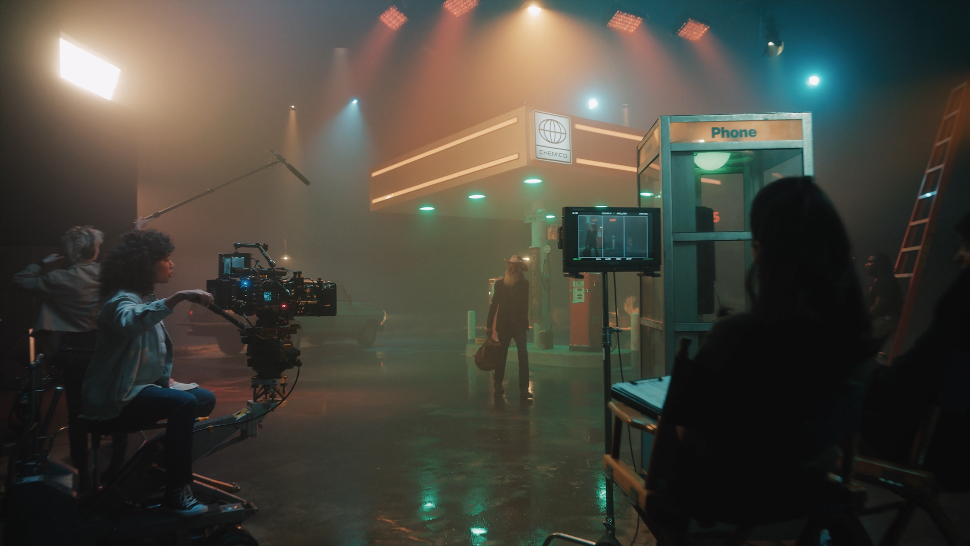 Film crew on set at a gas station, with a camera operator, director, and lighting equipment, under atmospheric lighting.