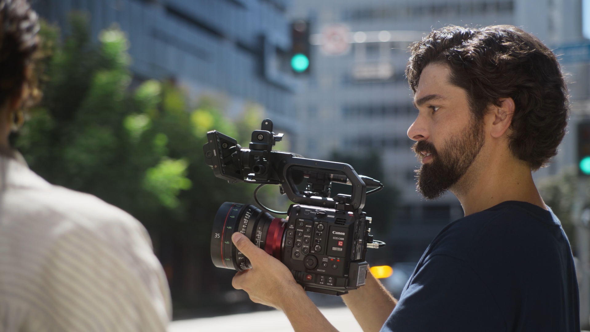 A man with dark, curly hair and a beard filming with a professional camera on a city street, with green traffic light visible in the background.
