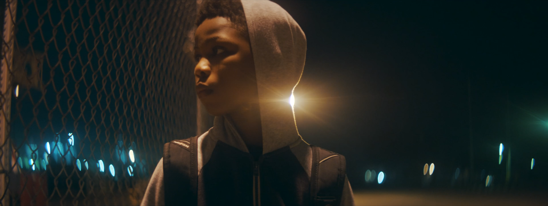 A young person wearing a hoodie and backpack standing near a chain-link fence at night, illuminated by streetlights in the background.