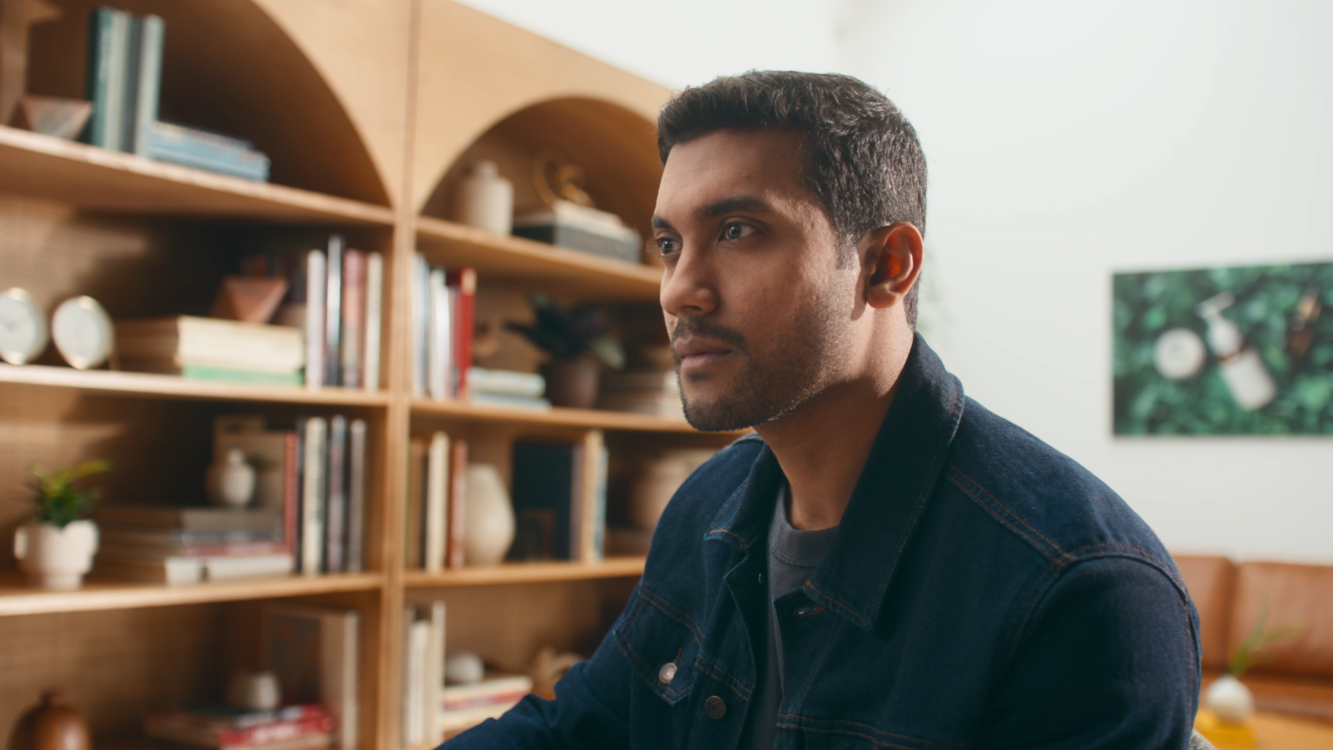 A man with short dark hair and a beard sitting indoors near a wooden bookshelf, looking to the side with a neutral expression.