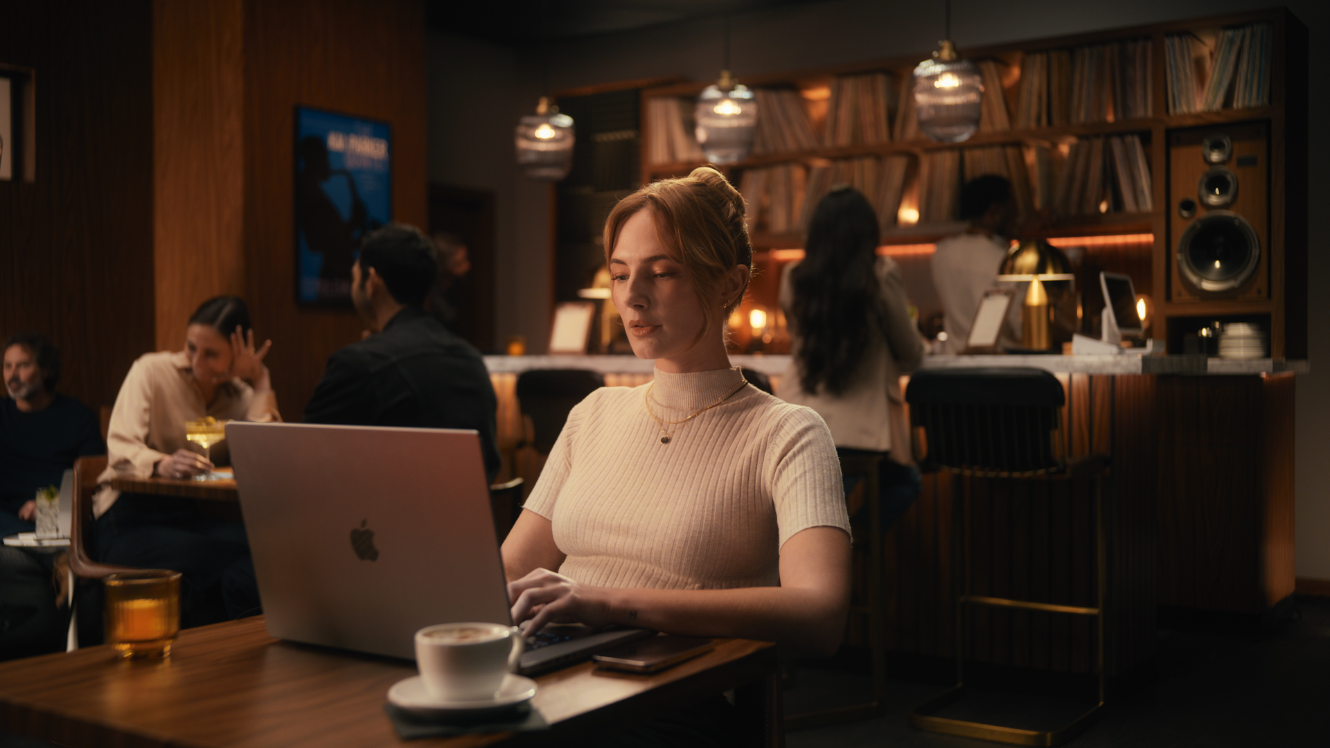 A woman with red hair working on a MacBook laptop in a cozy, dimly lit café or bar. She is wearing a beige ribbed turtleneck and gold jewelry. In the background, there are other people socializing, a bar counter with glasses and a large speaker, and a bookshelf filled with books, creating a warm atmosphere.