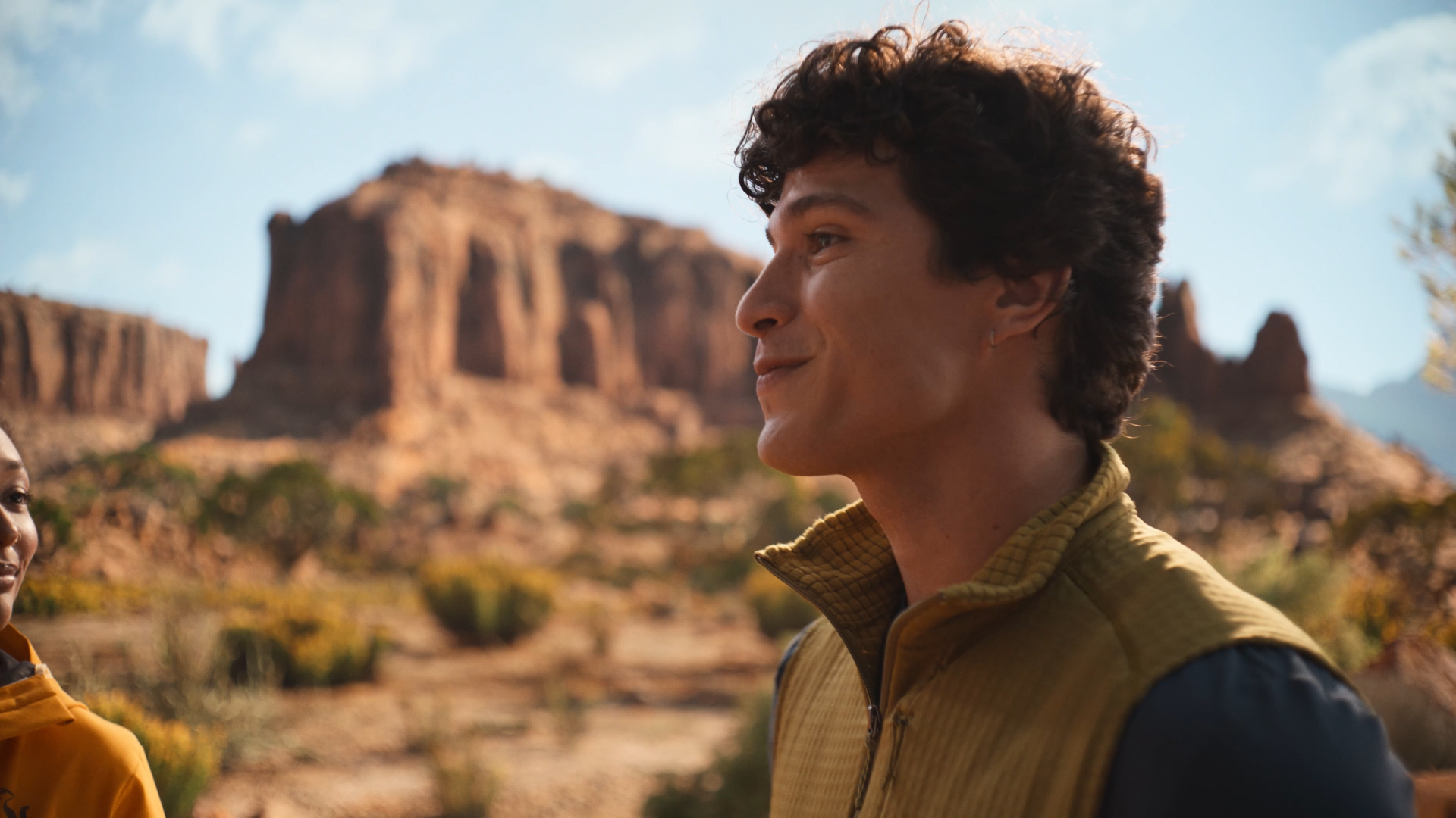 Side profile of a smiling young man with curly hair wearing a tan and black jacket, standing outdoors in a desert landscape with large rock formations and blue sky in the background.