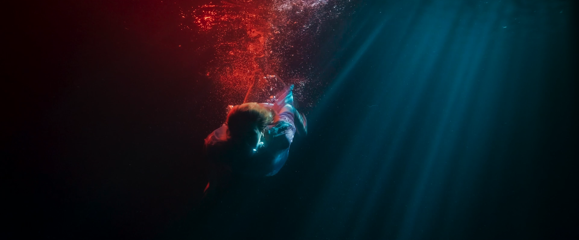 Person swimming underwater with light filtering through the water, bubbles surrounding them, and red and blue lighting effects.