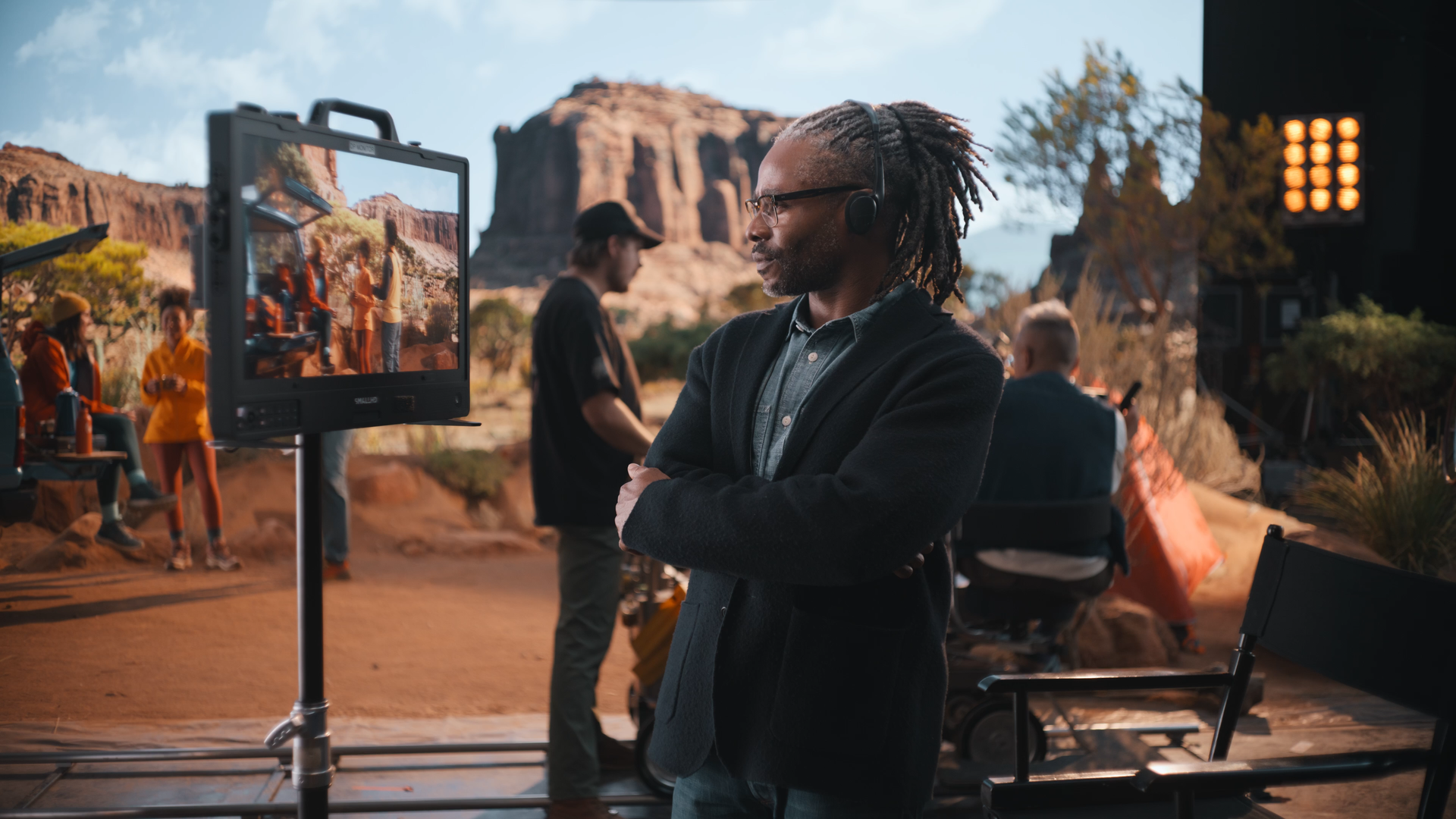 A film crew working on a desert set with red rock formations, with a man with glasses and dreadlocks in the foreground wearing a headset.