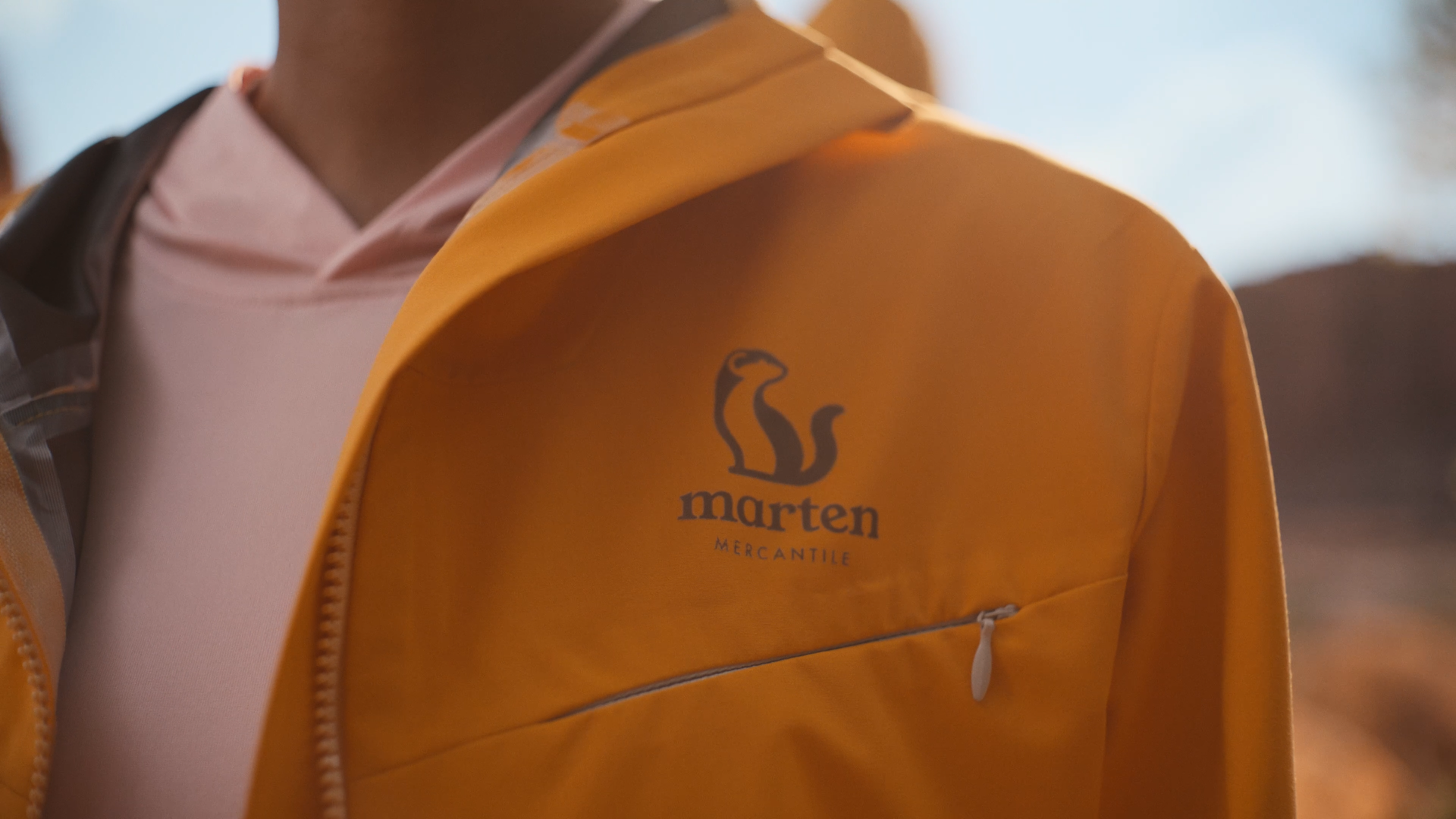 A person wearing a pink hoodie and a tan jacket with a logo and text. The logo depicts a stylized animal, possibly a lion or cat, with the words 'marten' and 'MERCANTILE' underneath.
