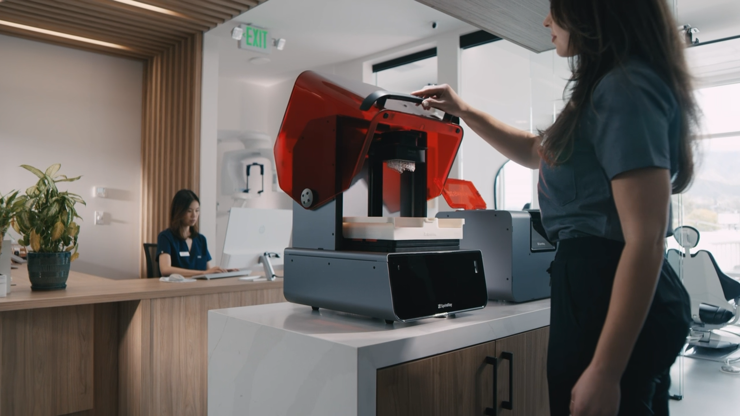 A woman in a gray shirt uses a 3D printer in a modern office or clinic setting, with a receptionist seated at a desk in the background, a potted plant, and large windows letting in natural light.