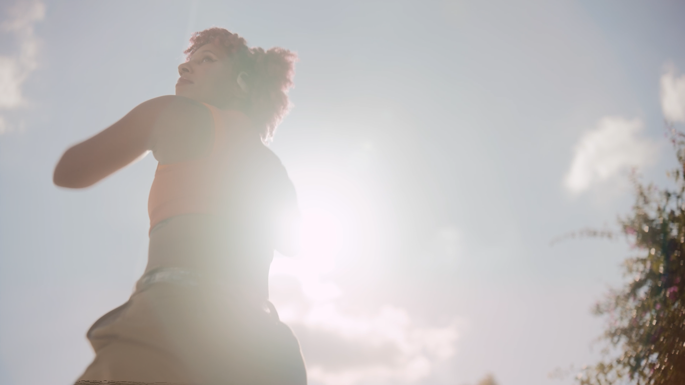 A person with curly hair is outside, with the sun shining behind them, creating a backlit silhouette against a partly cloudy sky.
