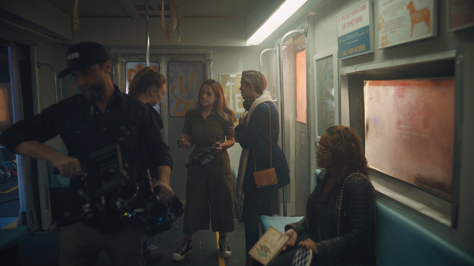 Four women and one man inside a train, with one woman sitting and holding a book, and the others standing and talking, the man adjusting camera equipment.