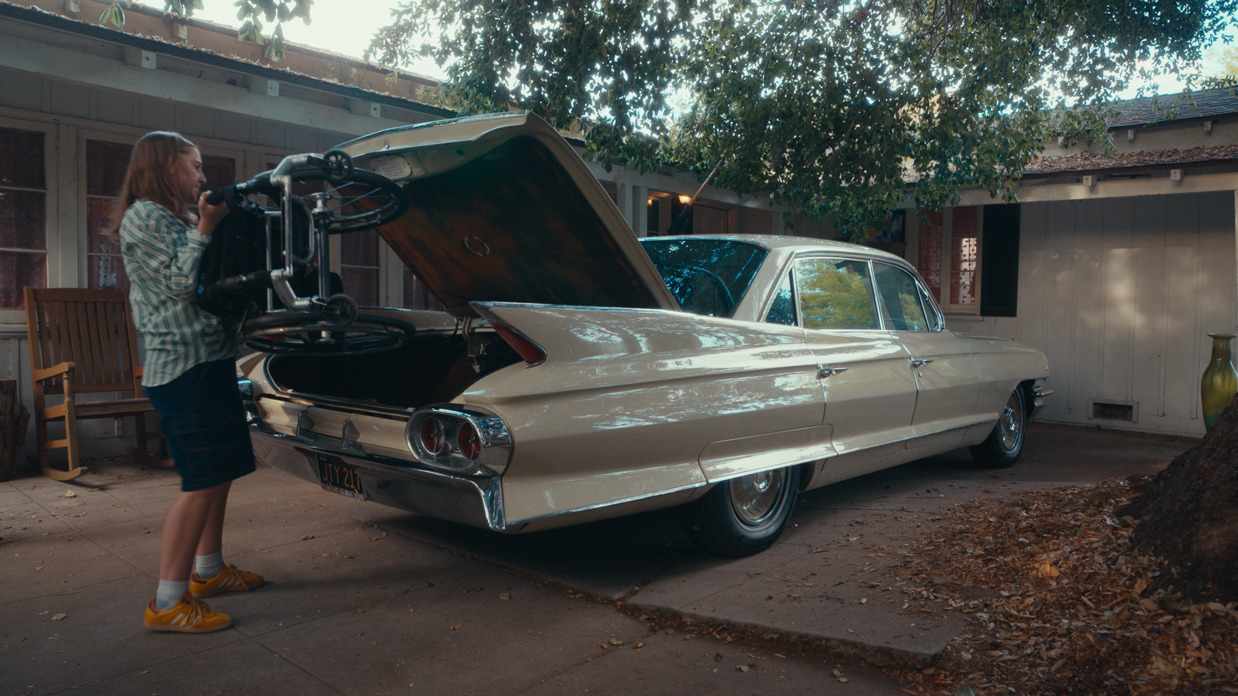 A young girl with red hair, wearing a plaid shirt, black shorts, and yellow sneakers, opening the trunk of a vintage beige car, possibly a Cadillac, parked in a backyard with trees, a house, and patio furniture.