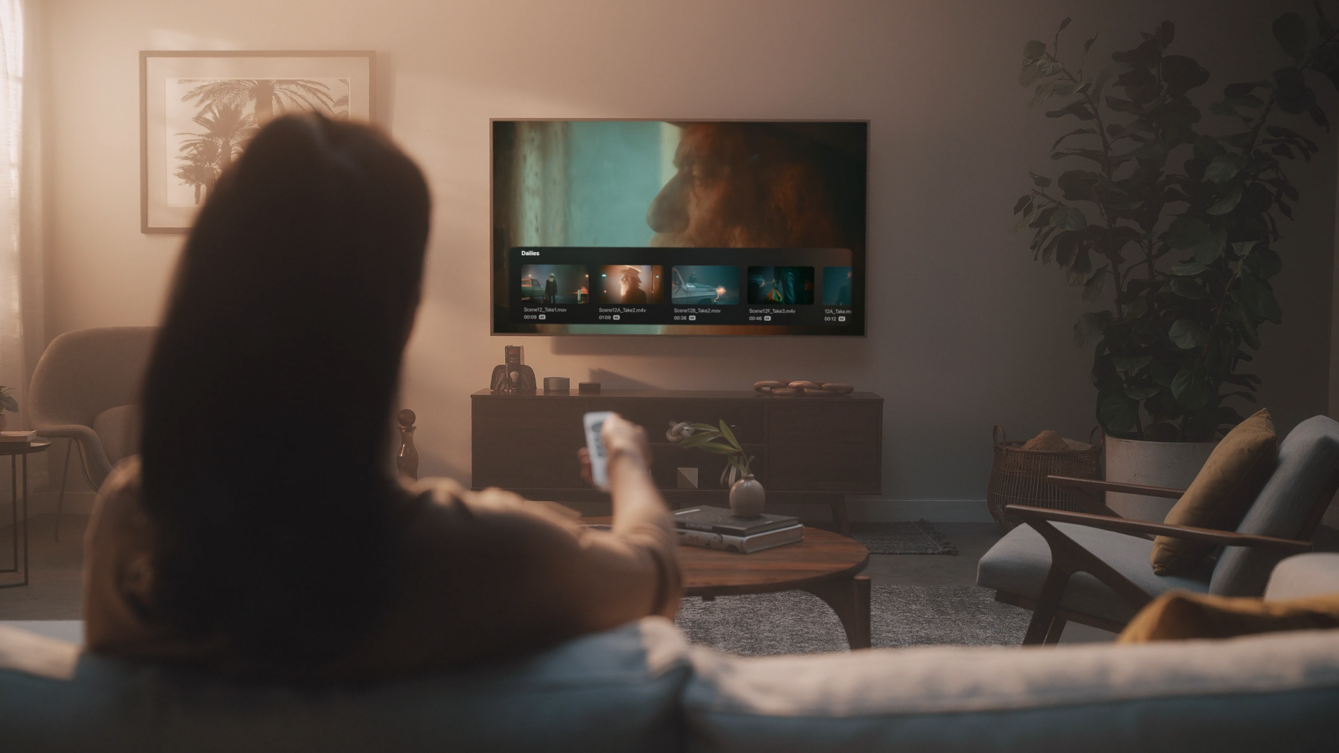 Person sitting on a sofa at home, watching television with a remote in hand. The TV shows a close-up of a person's face and a film scene. The room has a framed picture of palm trees on the wall, a plant in a pot, and a coffee table with magazines and decorative items. The space appears cozy and modern.