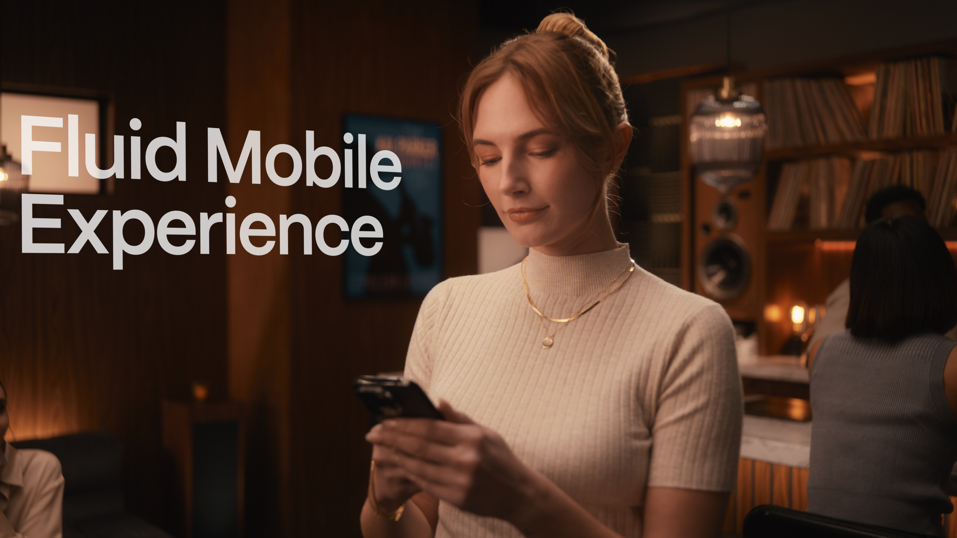 A woman with red hair in a beige turtleneck looks at her smartphone inside a cozy, dimly lit room with wooden walls and bookshelves.