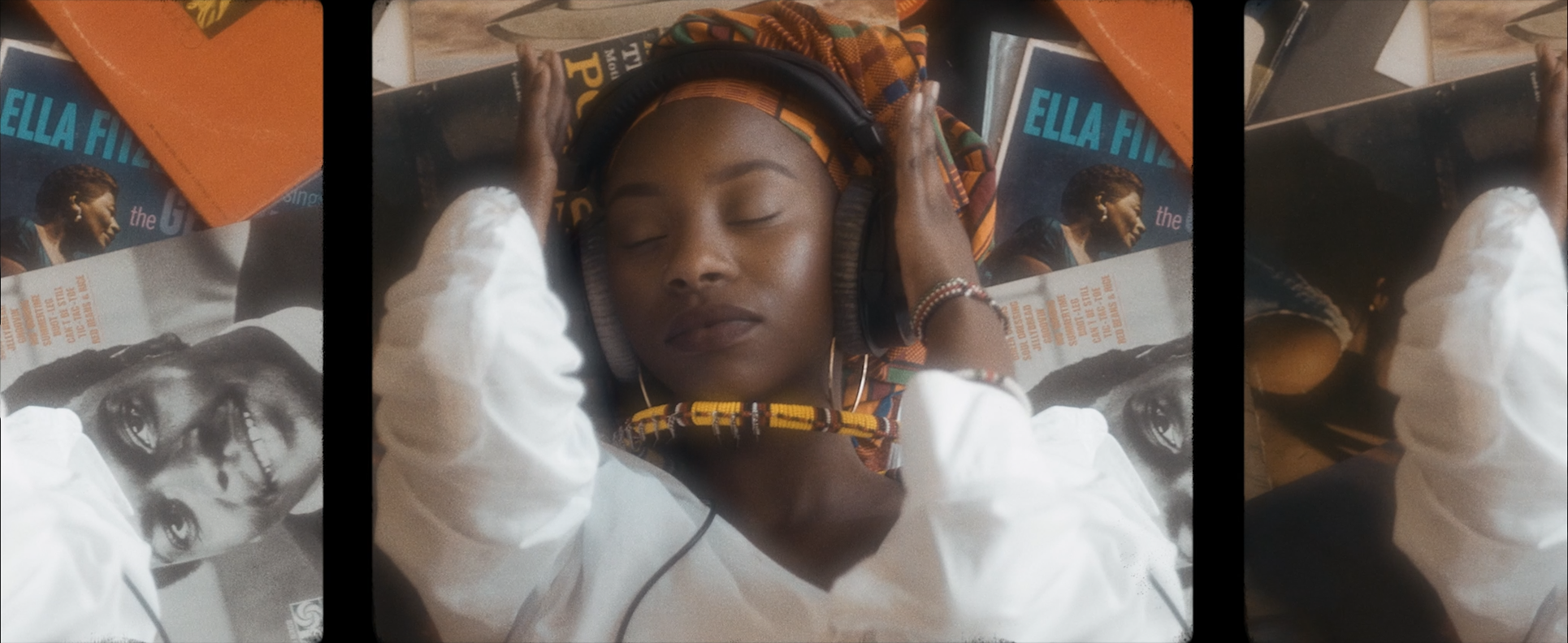 A young woman with closed eyes, wearing a colorful headwrap and large headphones, is resting her head on a table. Surrounding her are several magazines, including one featuring a smiling woman. She is wearing a white blouse and a beaded bracelet.