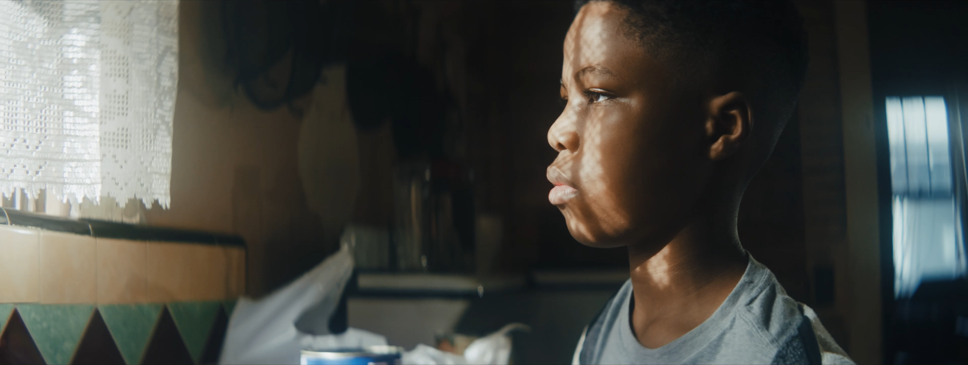 Profile of a young boy with short hair looking outside a window with sunlight casting shadows on his face.