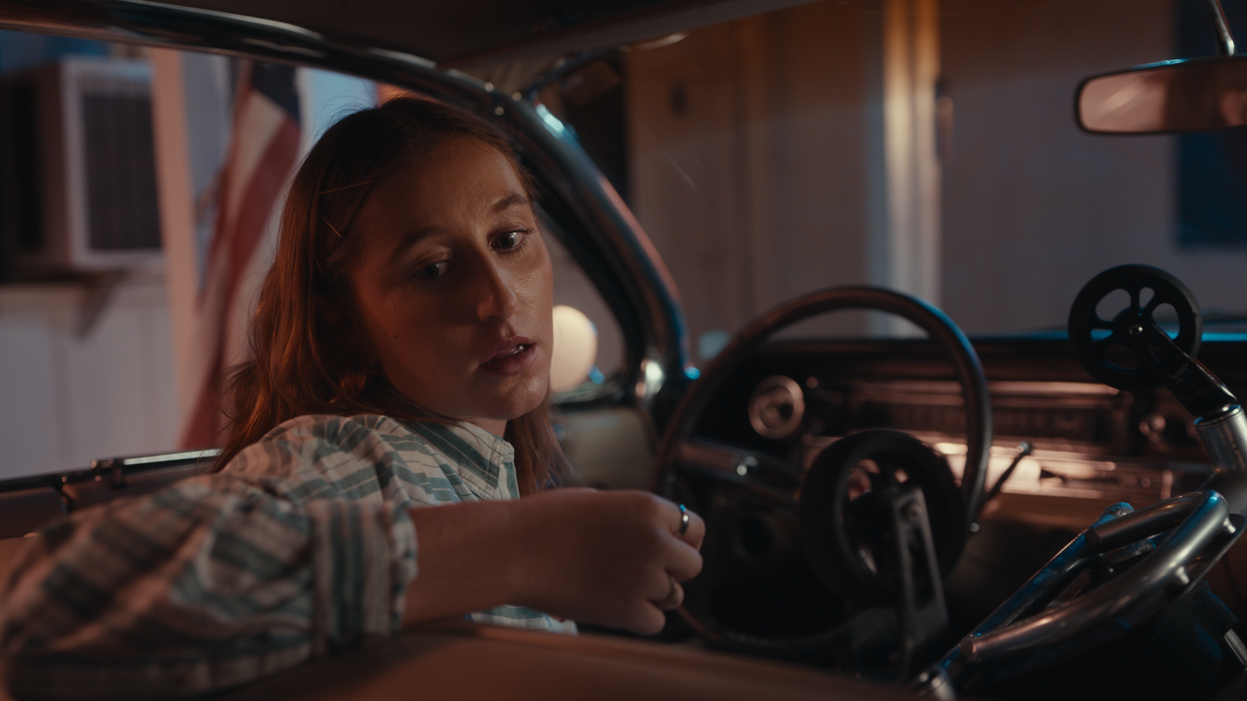 A young woman with long red hair and a plaid shirt sitting in the driver's seat of a vintage car.