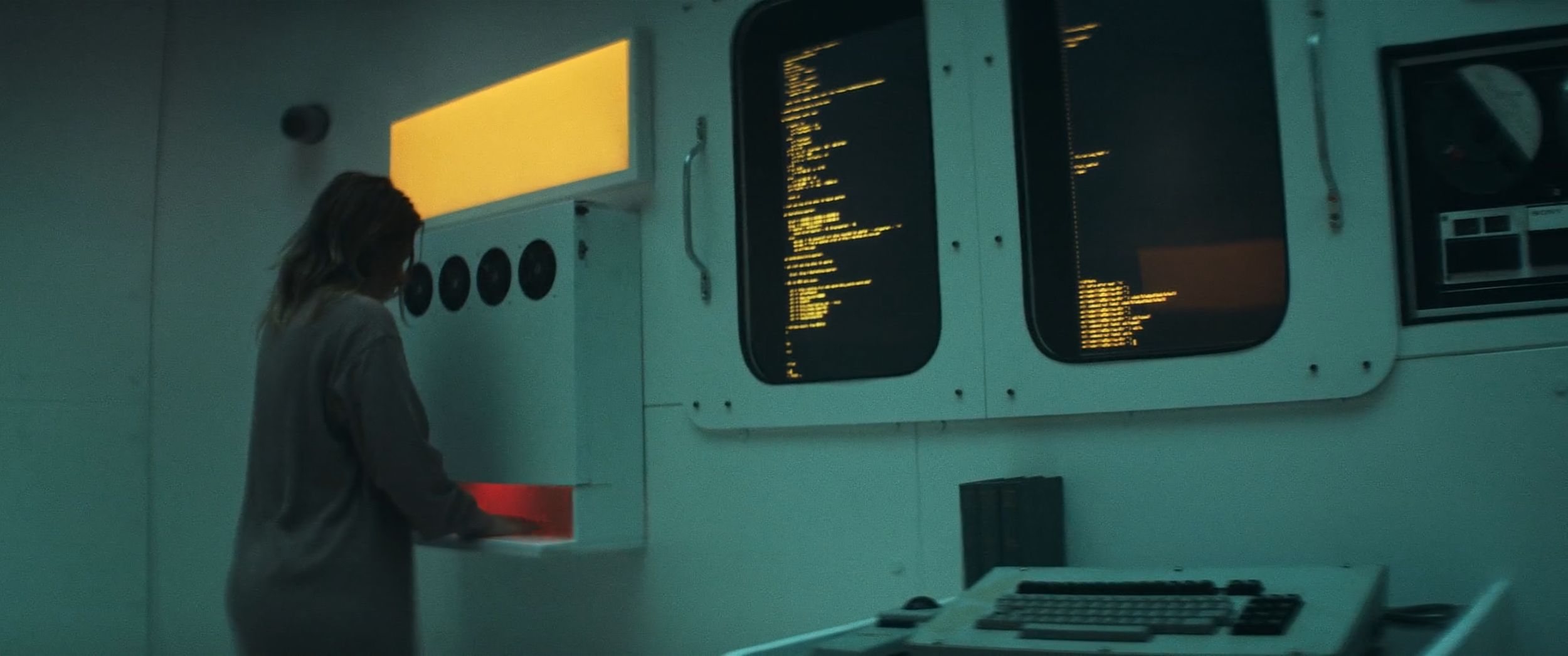 A woman in a gray hoodie stands in a room with a computer monitor and multiple screens displaying lines of code, suggesting a high-tech or control room environment.