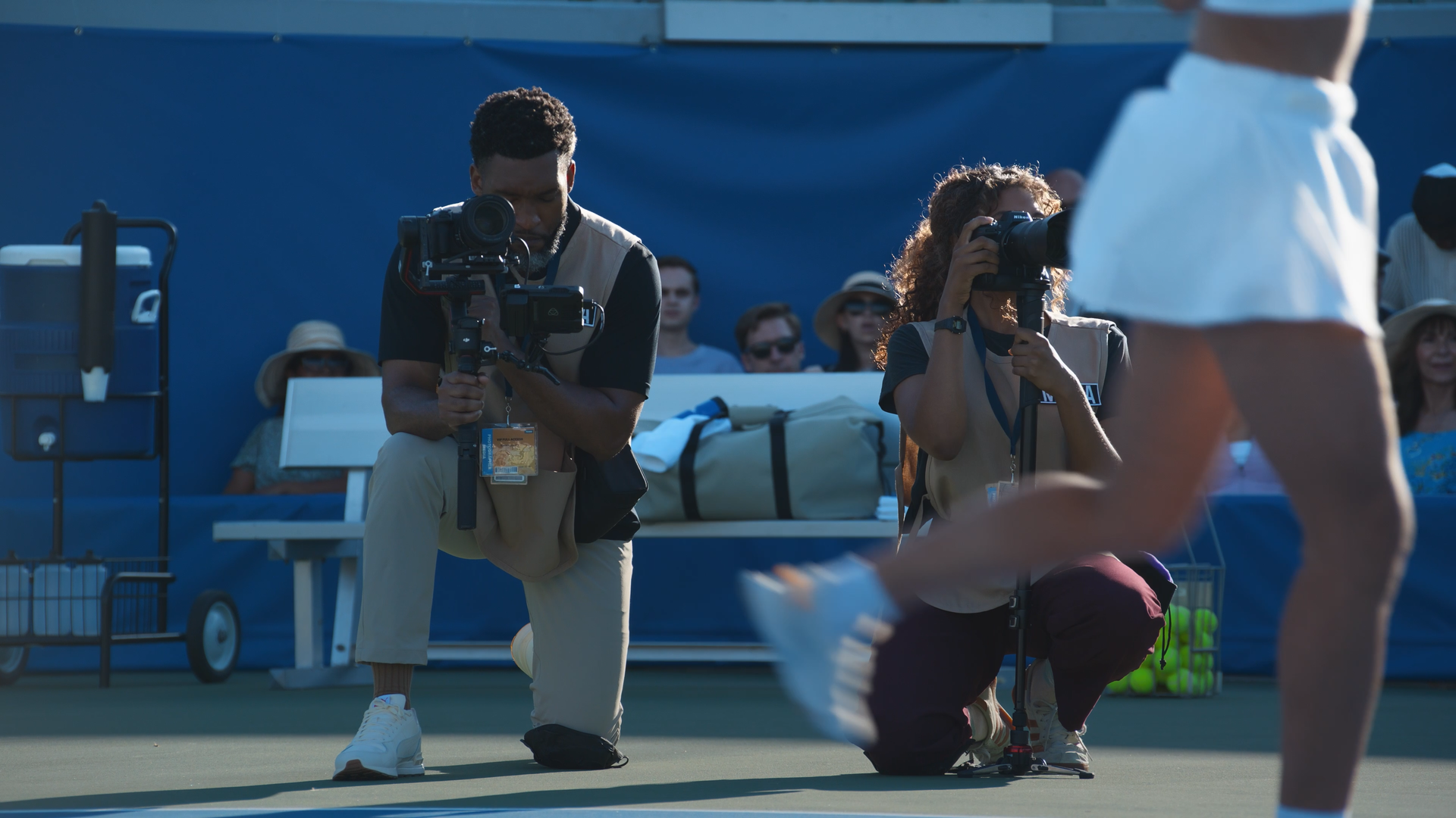 Photographers capturing a moment at a tennis event with a group of people sitting behind them.